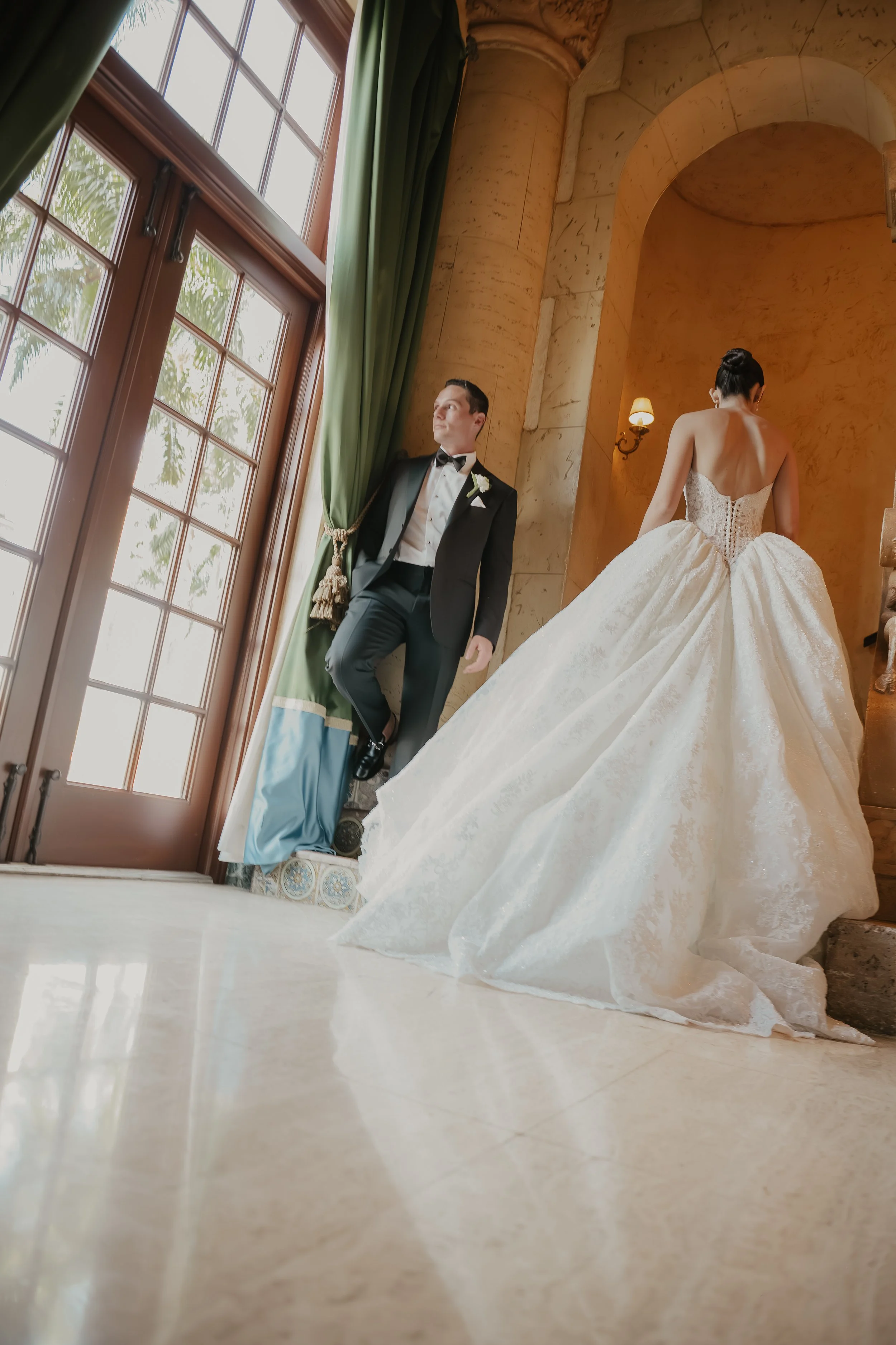 A bride in a white wedding gown with her back to the camera and a groom in a tuxedo standing beside her, near a large window with green curtains in a warmly lit room.