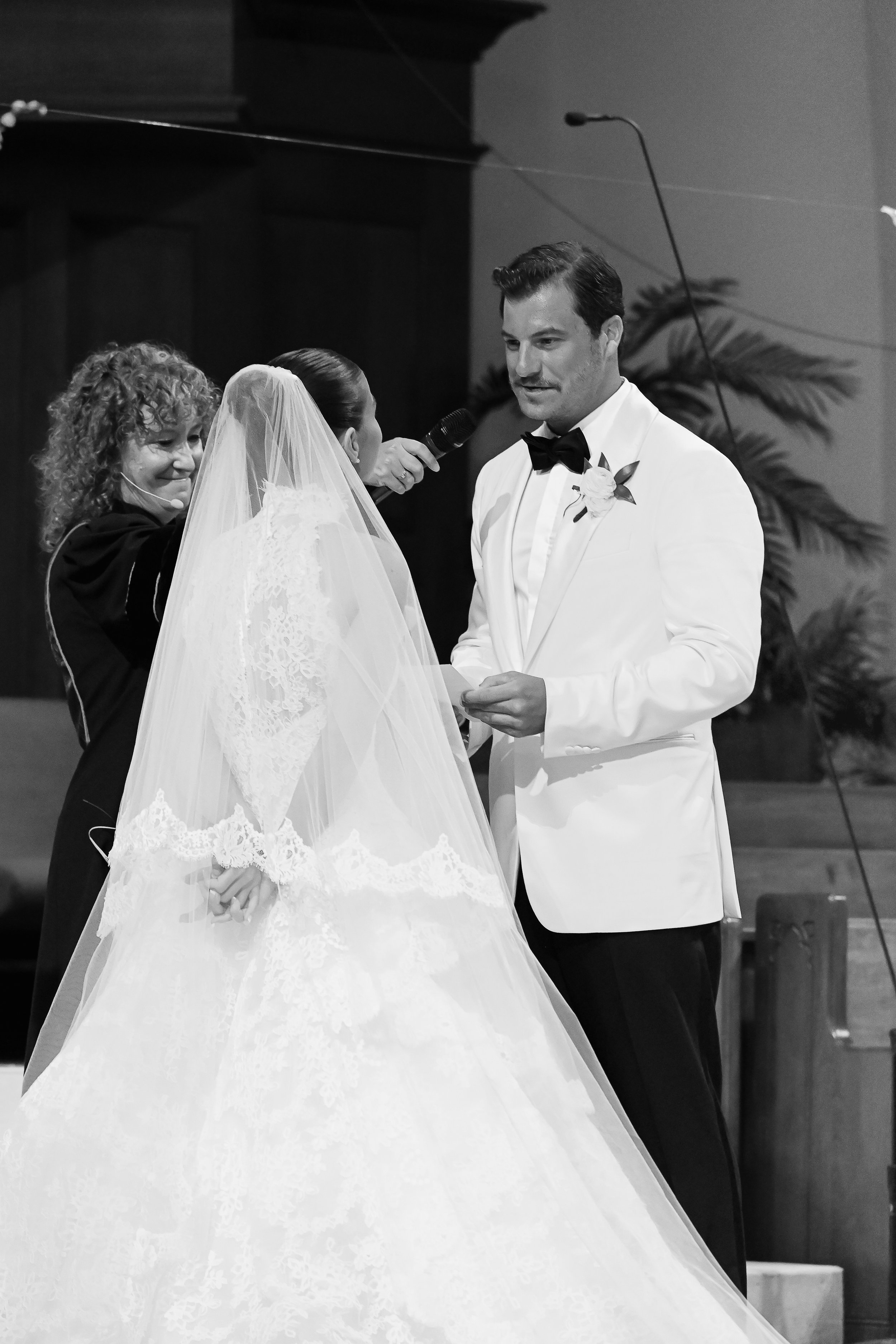A black and white photo of a wedding ceremony where a groom in a tuxedo and a bride in a wedding dress are holding hands and facing each other, with a woman officiant speaking into a microphone.