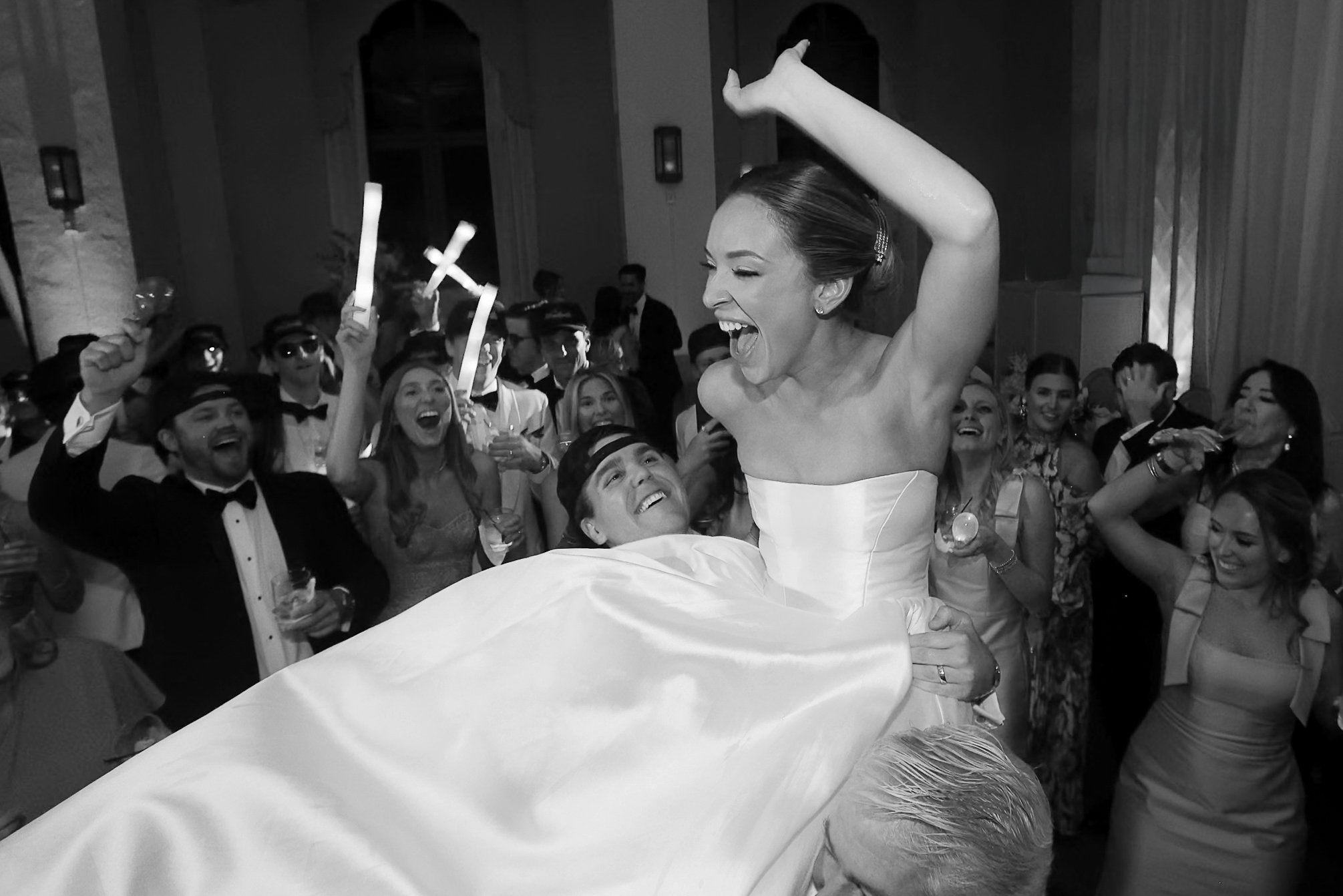 A joyful bride being lifted by a groom amidst a crowd of well-dressed wedding guests at a reception, celebrating with smiles and cheers.
