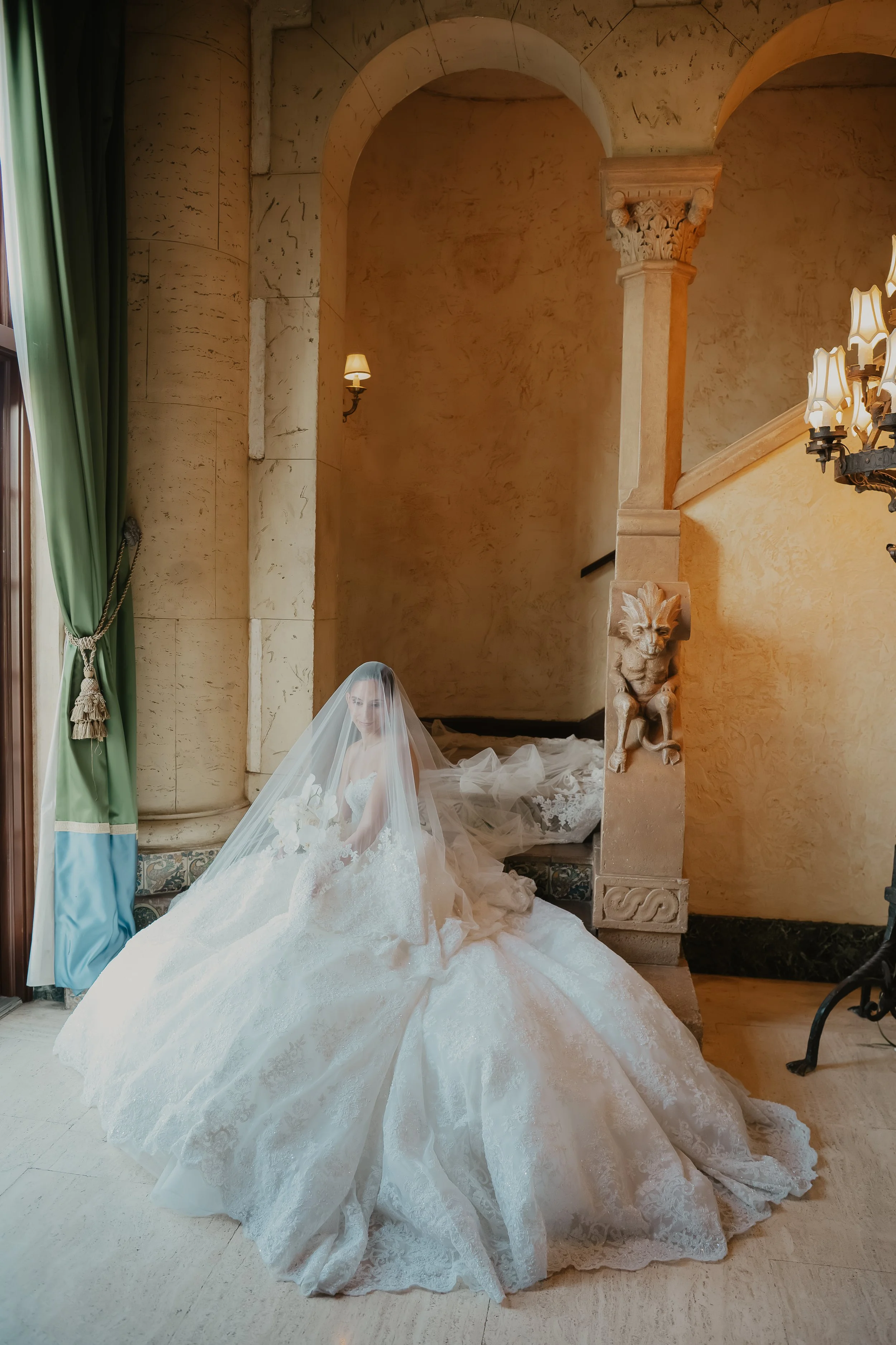 A bride in a white wedding gown with a long train, standing in an elegant, classical interior with ornate columns, wall sconces, a chandelier, and green curtains. She is wearing a veil and holding a bouquet.