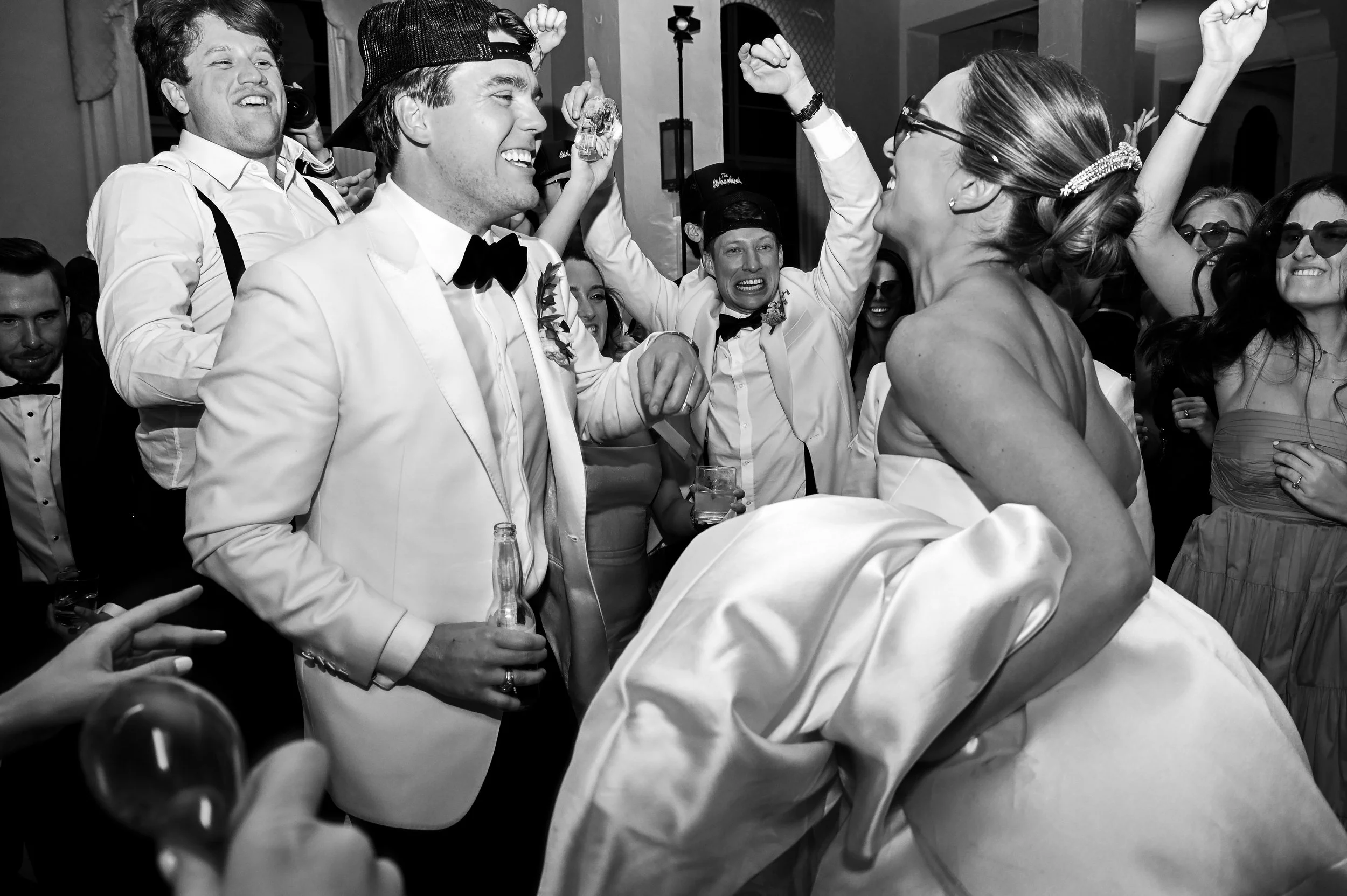 Black and white photo of people at a wedding celebration. Men in tuxedos and women in dresses, smiling and dancing, with some raising their arms in excitement.