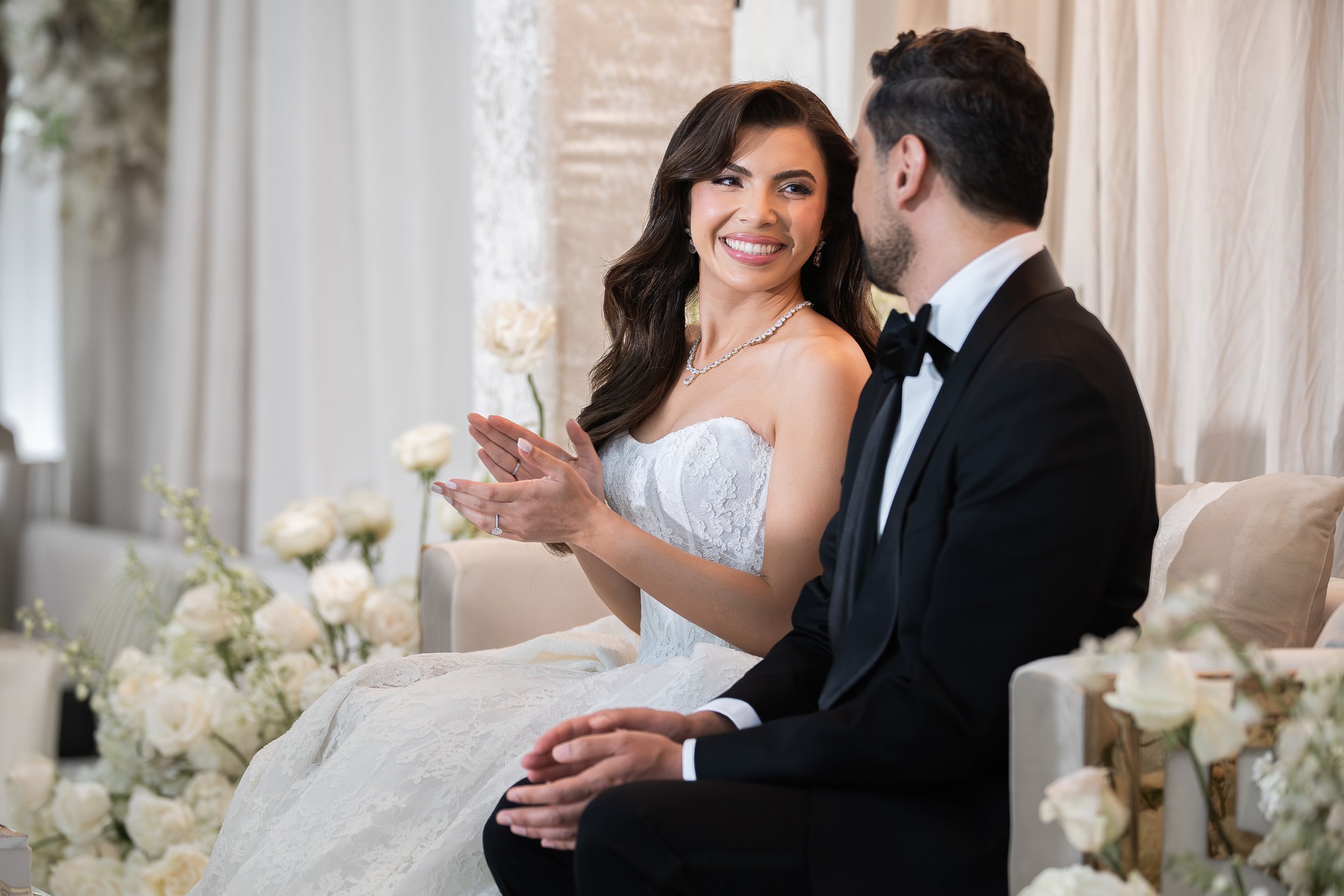 A bride in a white wedding dress and a groom in a black tuxedo sitting side by side at their wedding ceremony. The bride is smiling and looking at the groom, with flowers and drapery in the background.