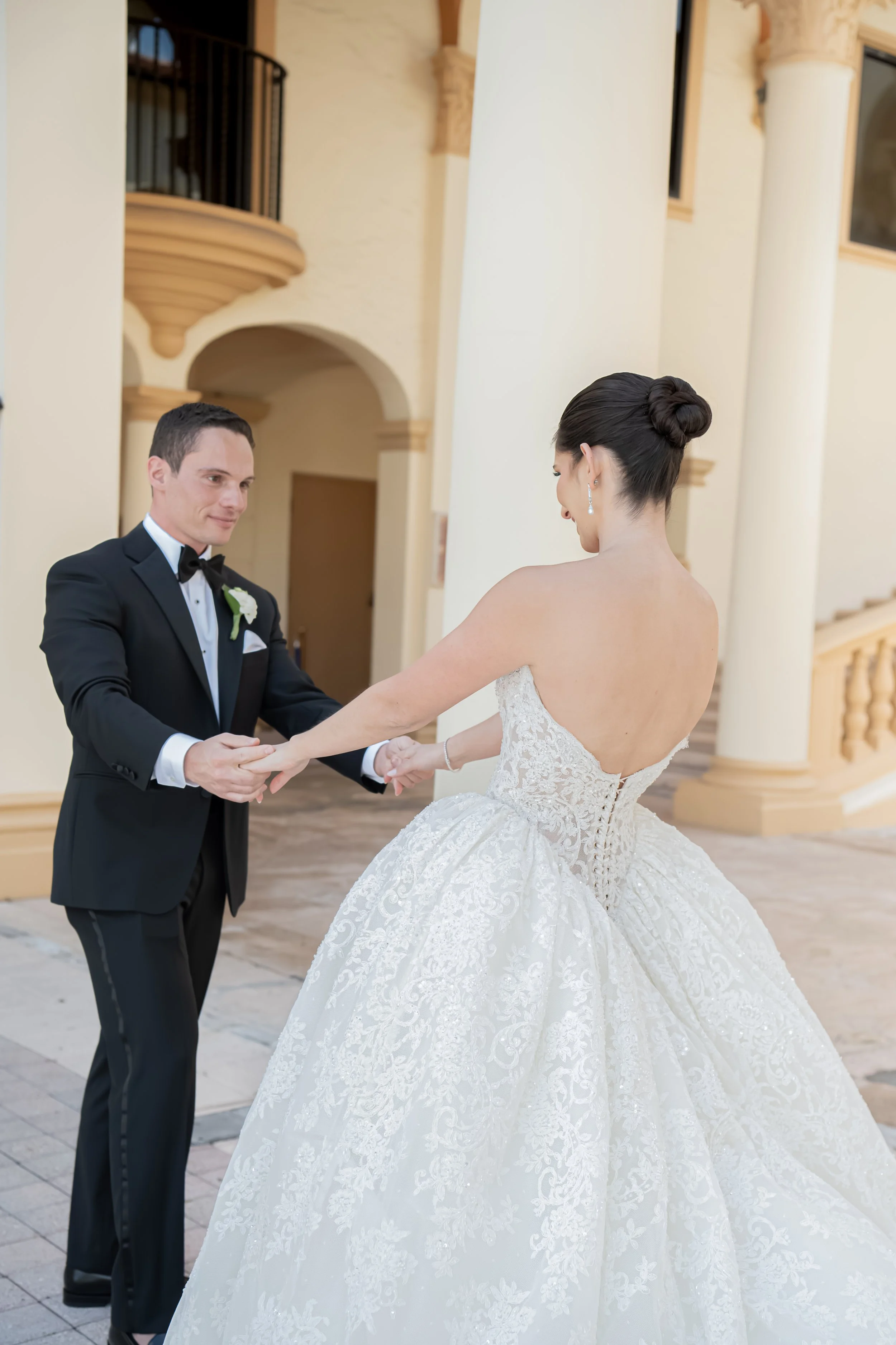 A bride and groom holding hands during their wedding ceremony indoors, with the bride in a white lace gown and the groom in a black tuxedo.