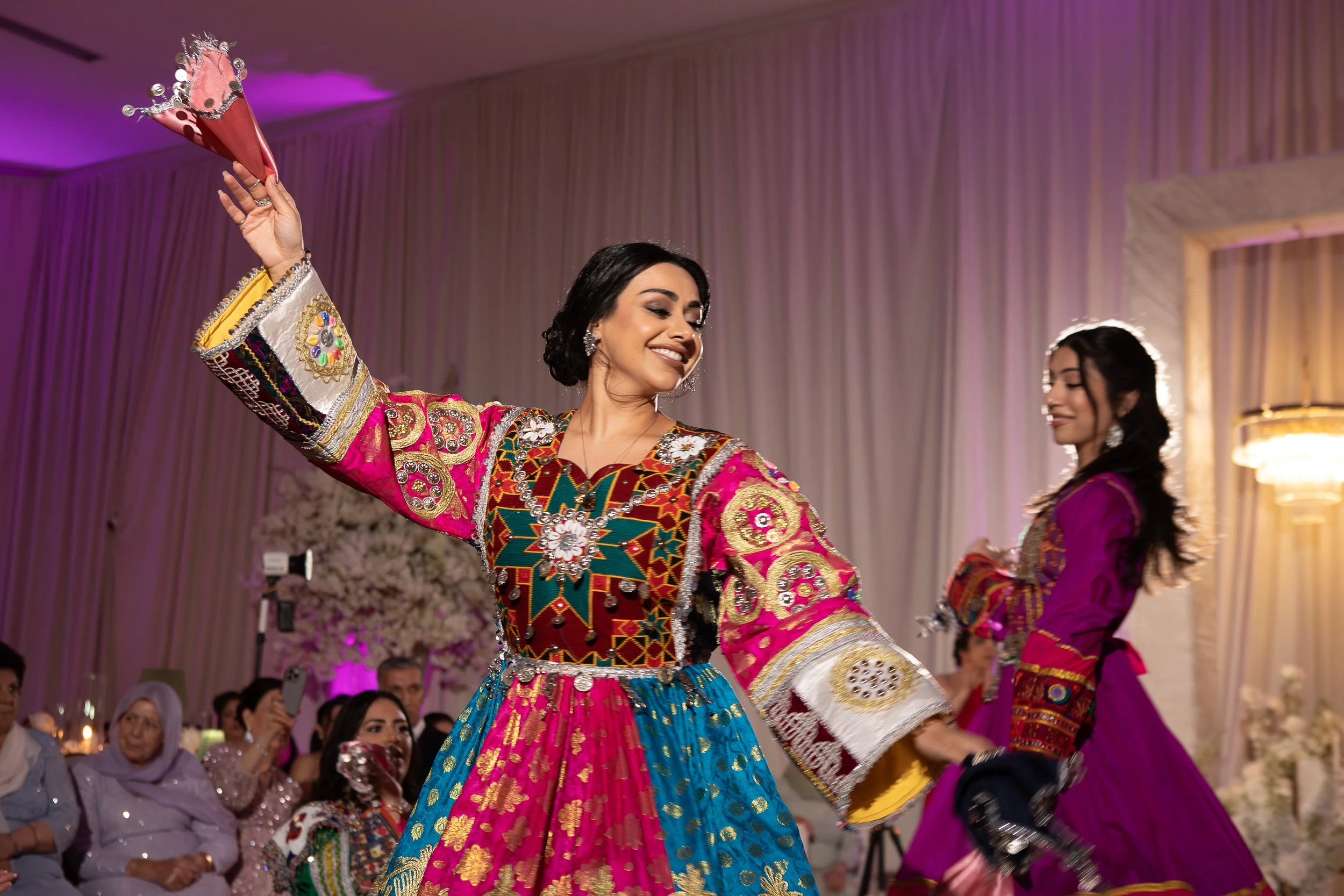 Two women dancing in colorful traditional dresses at an indoor celebration with a decorated background and seated audience.