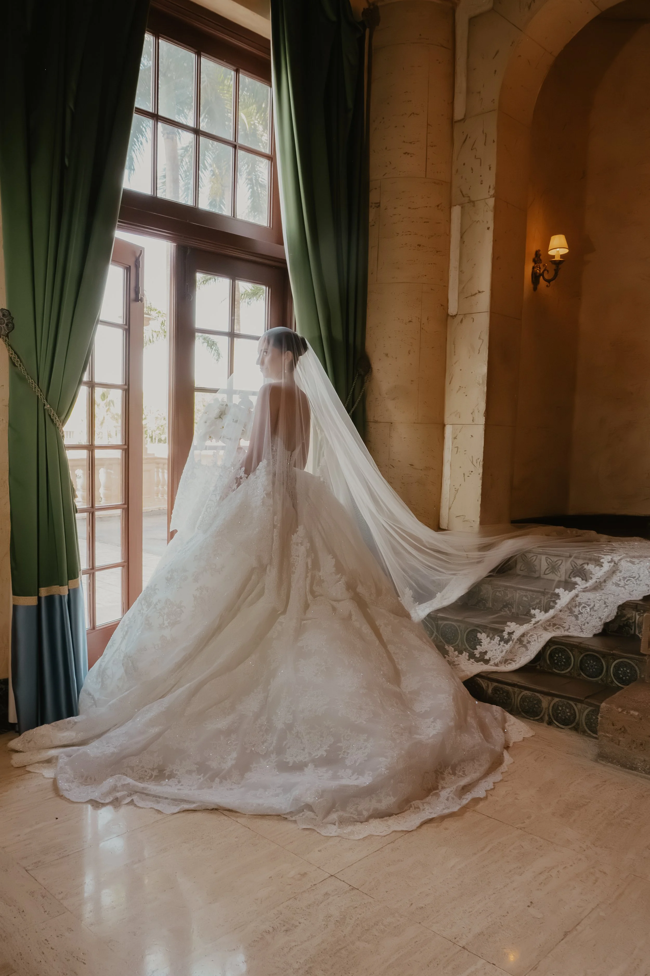 A bride in a wedding gown with a long train and veil standing near large window with green curtains, illuminated by natural light, in a room with beige marble walls and a wall-mounted light fixture.