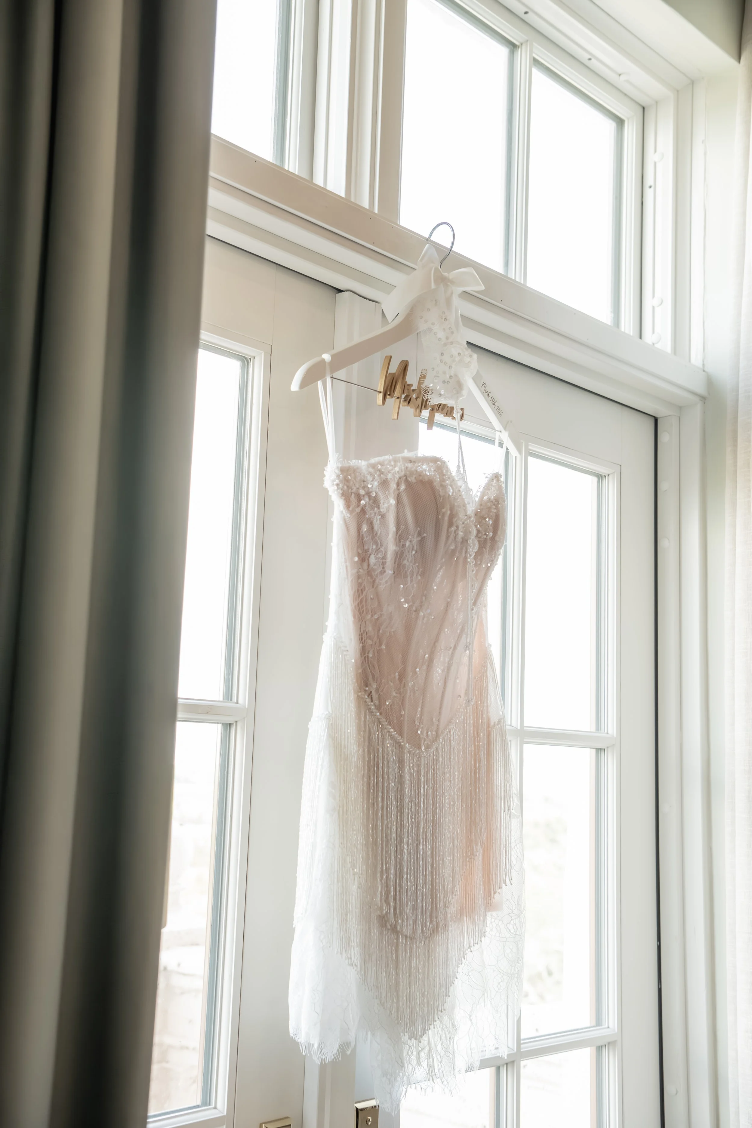 A white wedding dress hanging on a hanger inside a bright room, with sunlight coming through the windows.