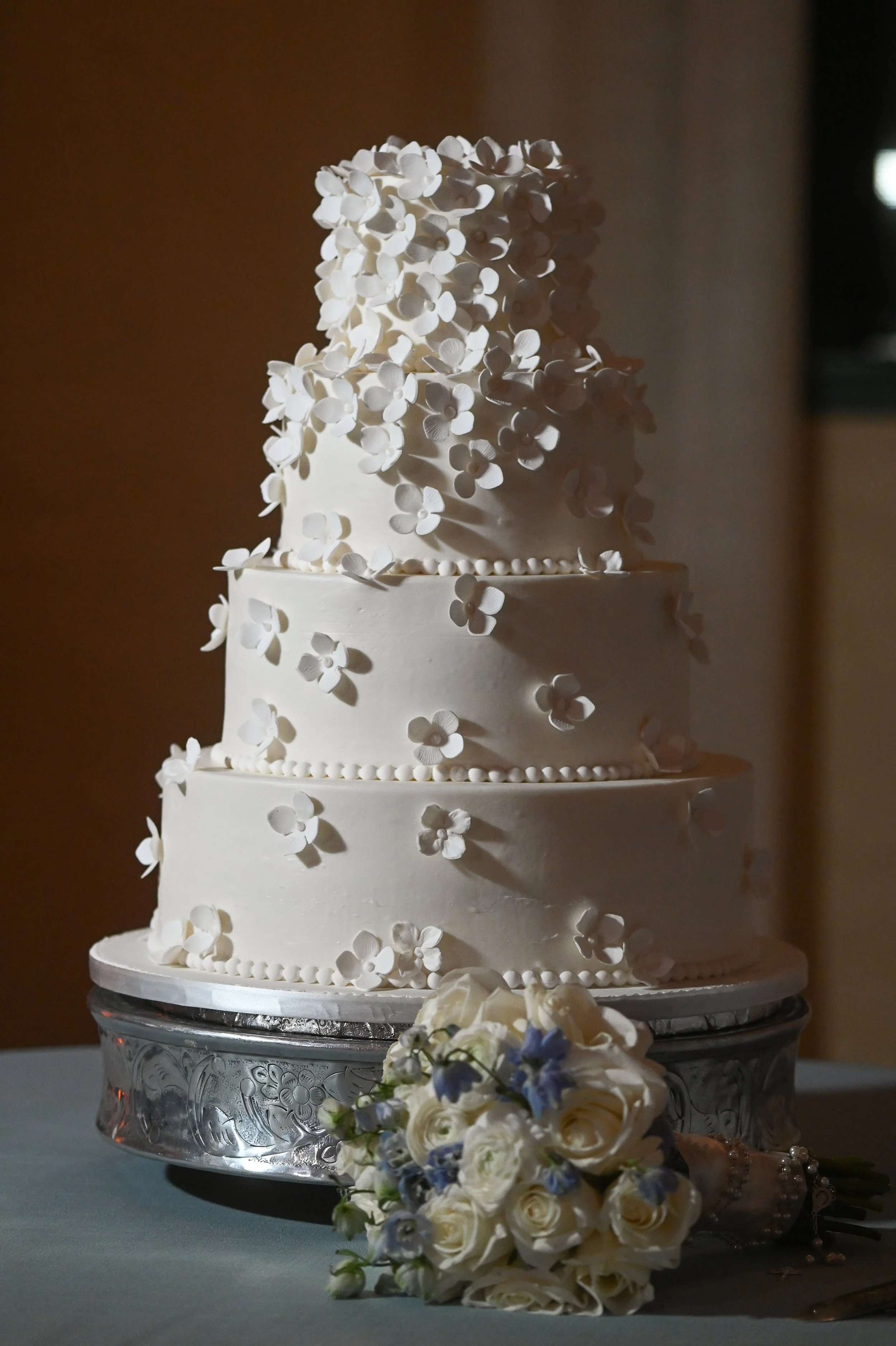 Three-tiered white wedding cake decorated with white and light purple flowers, placed on a decorative silver cake stand, with a bouquet of white and purple flowers and pearls in the foreground.
