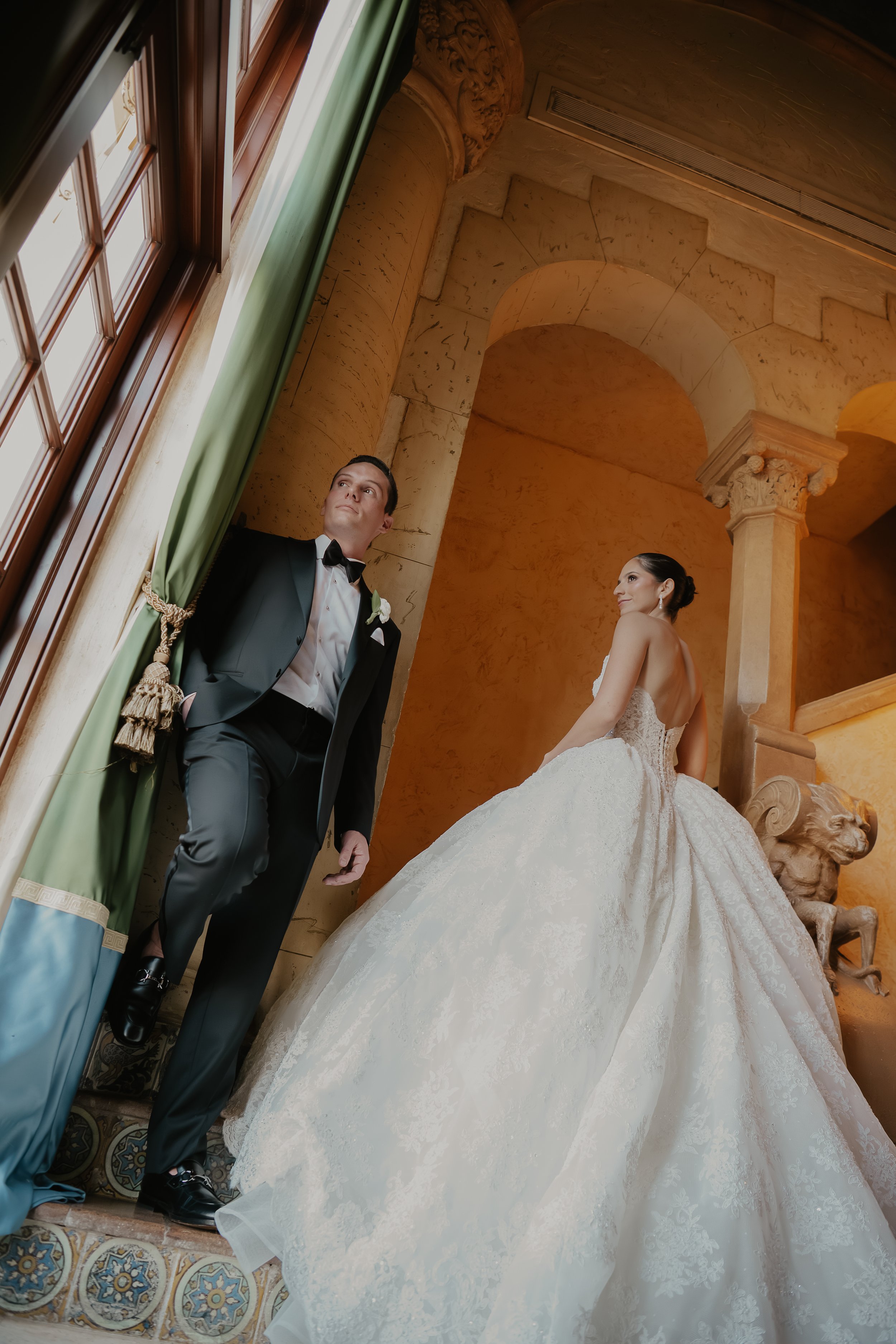 A bride and groom posing indoors with ornate architecture and a lion sculpture in the background. The groom wears a black tuxedo with a bow tie, and the bride wears a white wedding gown with lace details.