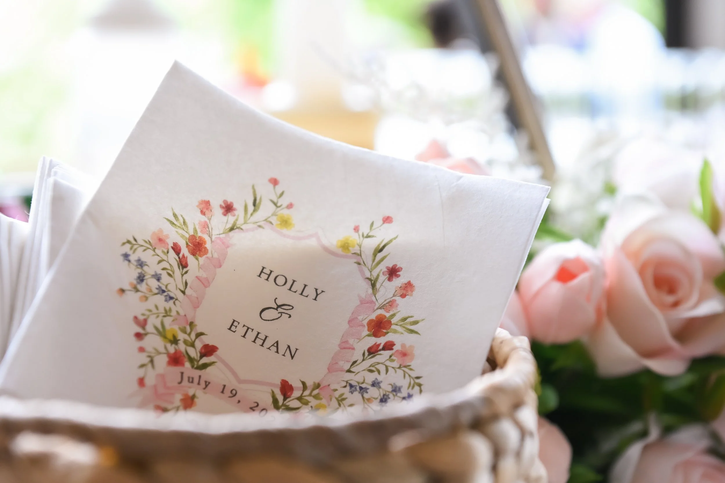Close-up of a wedding invitation card with floral designs, placed in a basket filled with pink roses, with blurred background.