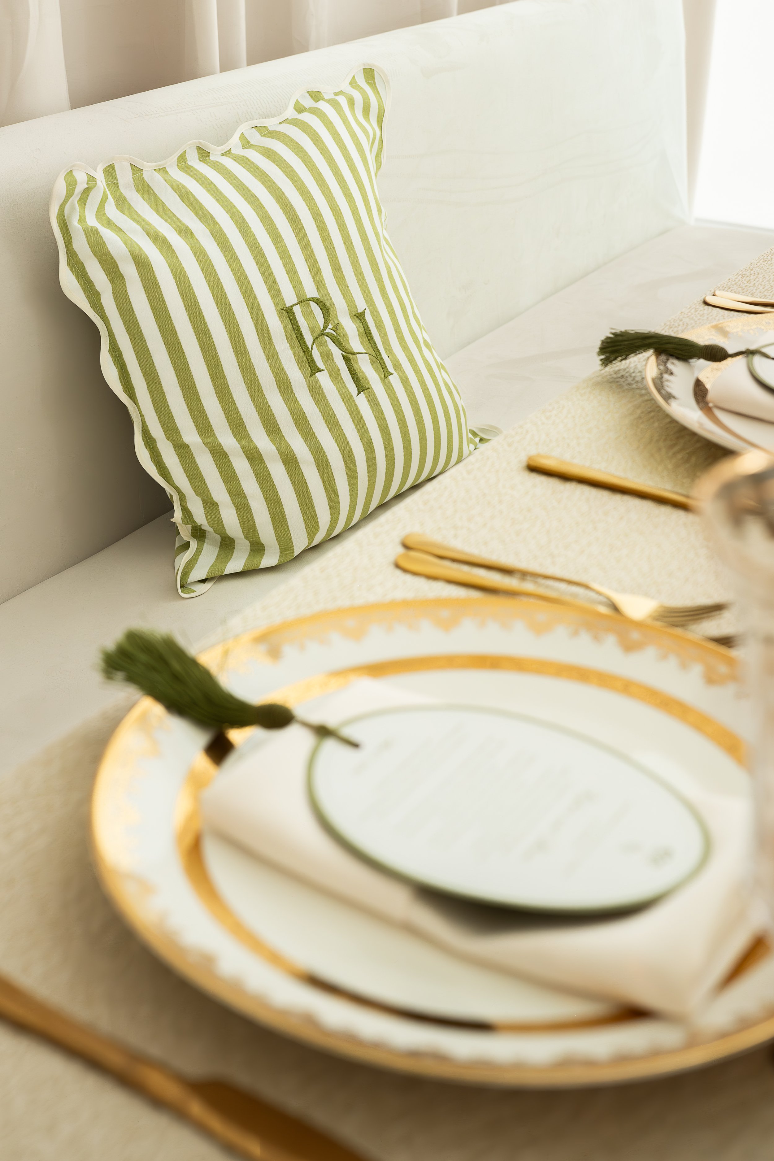 A green and white striped decorative pillow with a monogram letter R embroidered on it, placed on a white bed with a yellow and white table setting nearby.