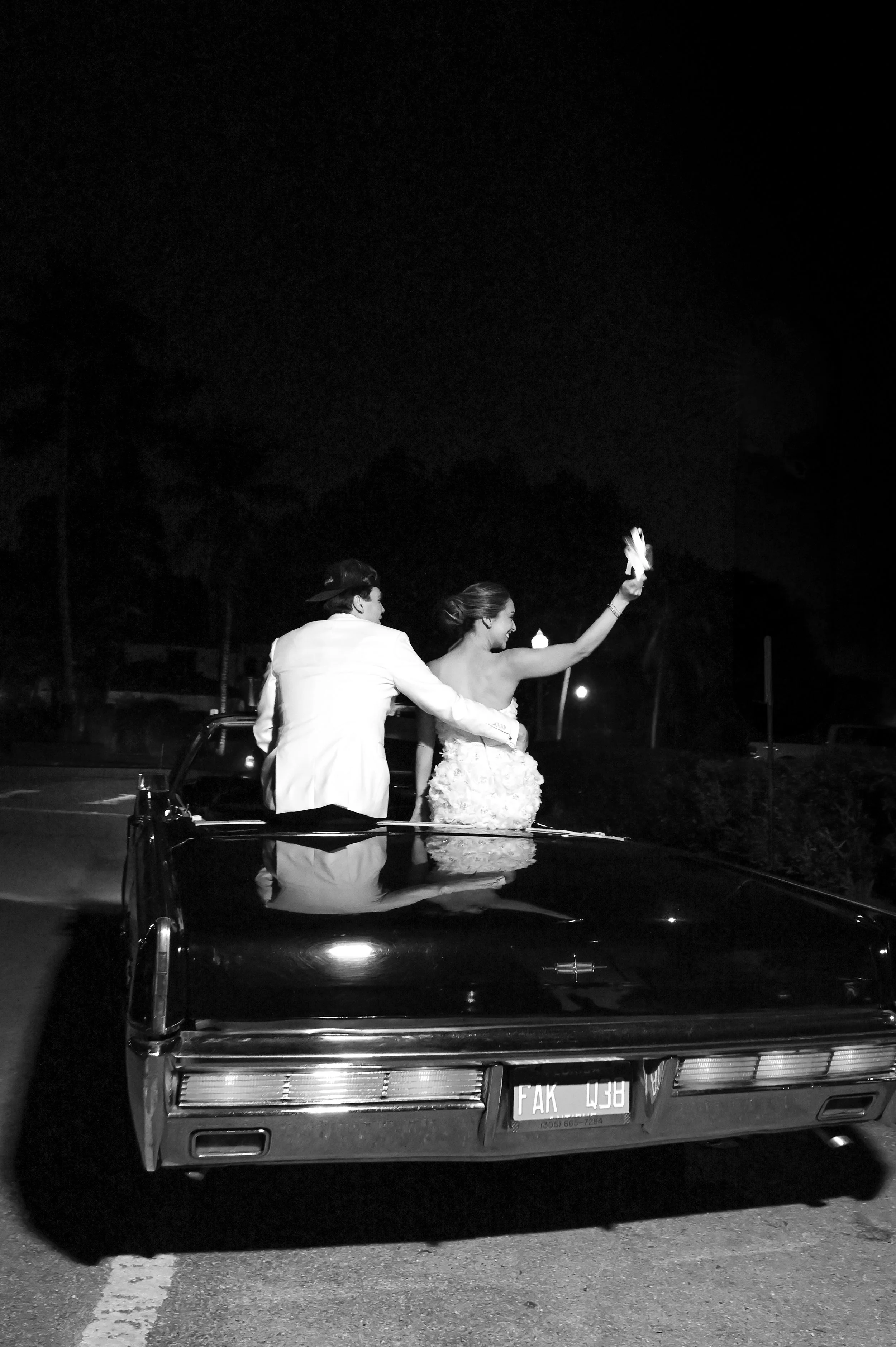 A black vintage car with a couple standing on it under the night sky. The woman is waving with a napkin, smiling, and wearing a wedding dress. The man is wearing a white suit and a hat, facing away from the camera.