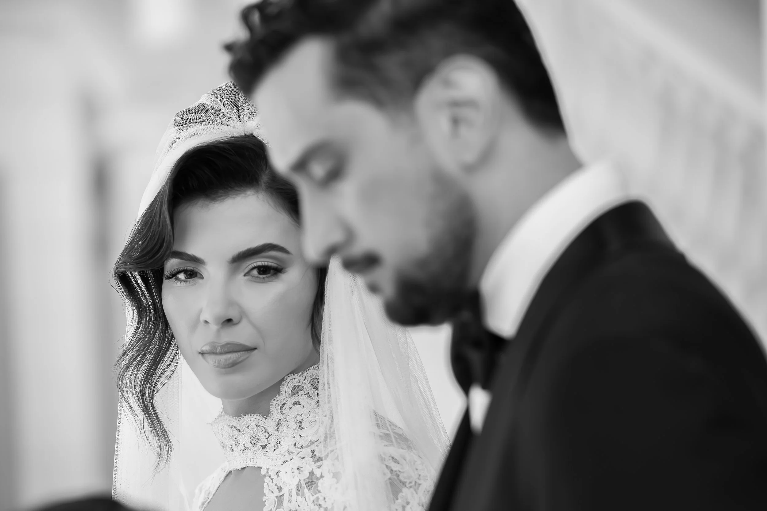 A bride and groom during a wedding ceremony, with the bride looking at the camera and the groom in profile, both dressed in wedding attire.