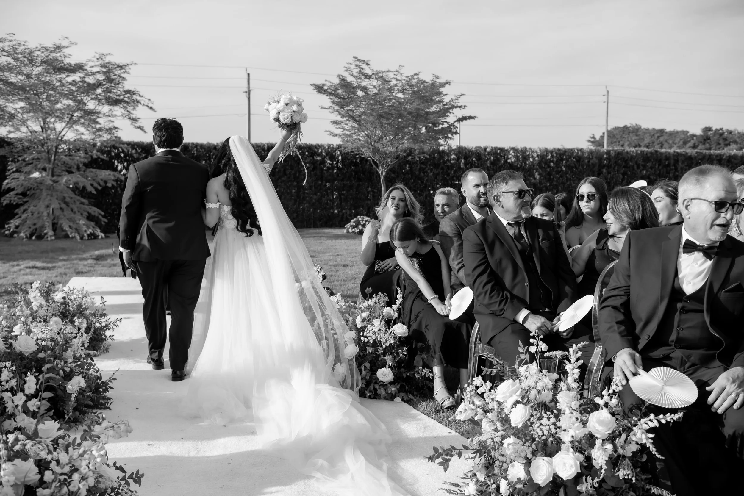 A black and white image of a wedding ceremony with the bride and groom standing at the altar, surrounded by seated guests outdoors.