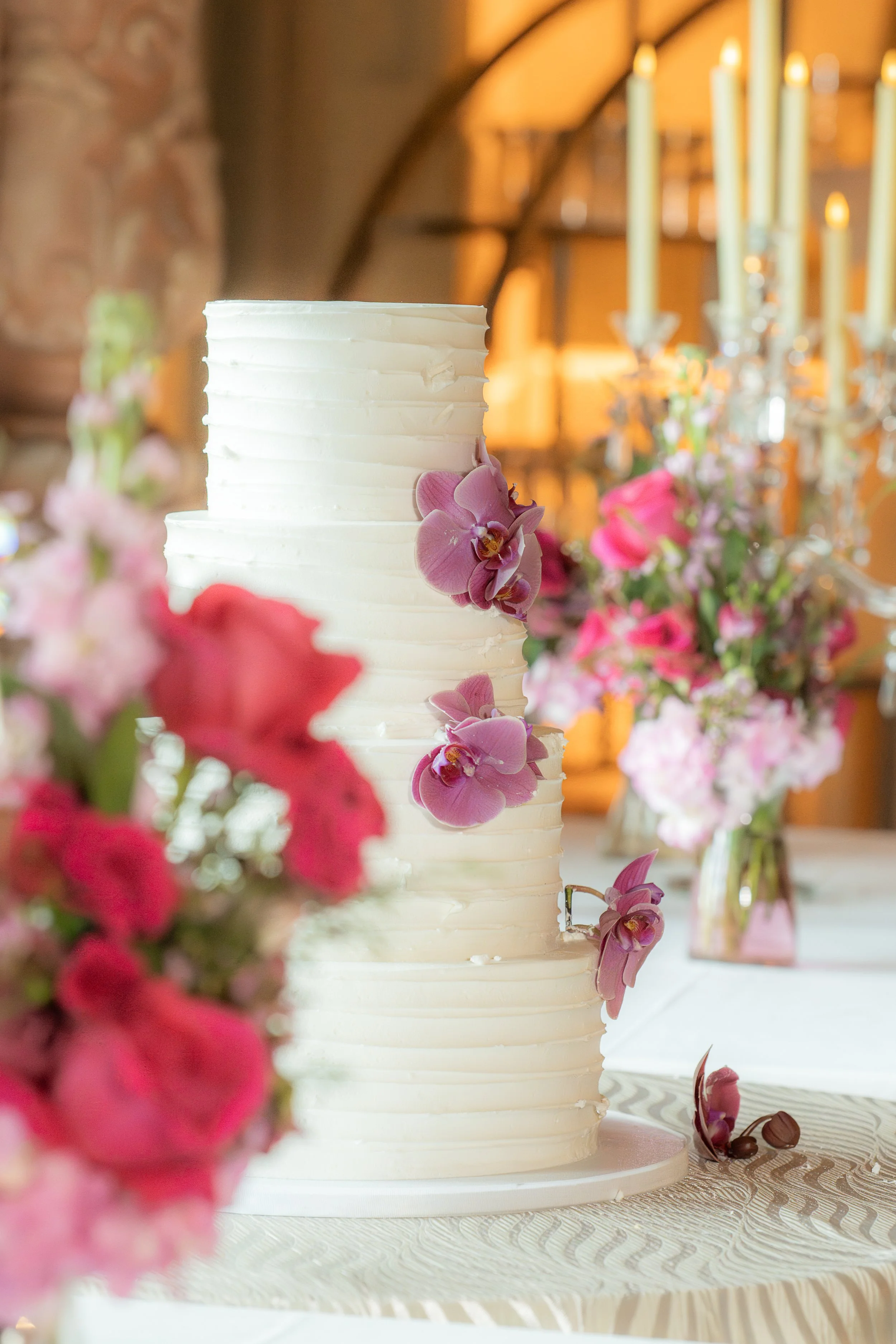 A white tiered wedding cake decorated with pink orchids, positioned in front of a floral arrangement and candle chandelier in an indoor wedding reception setting.