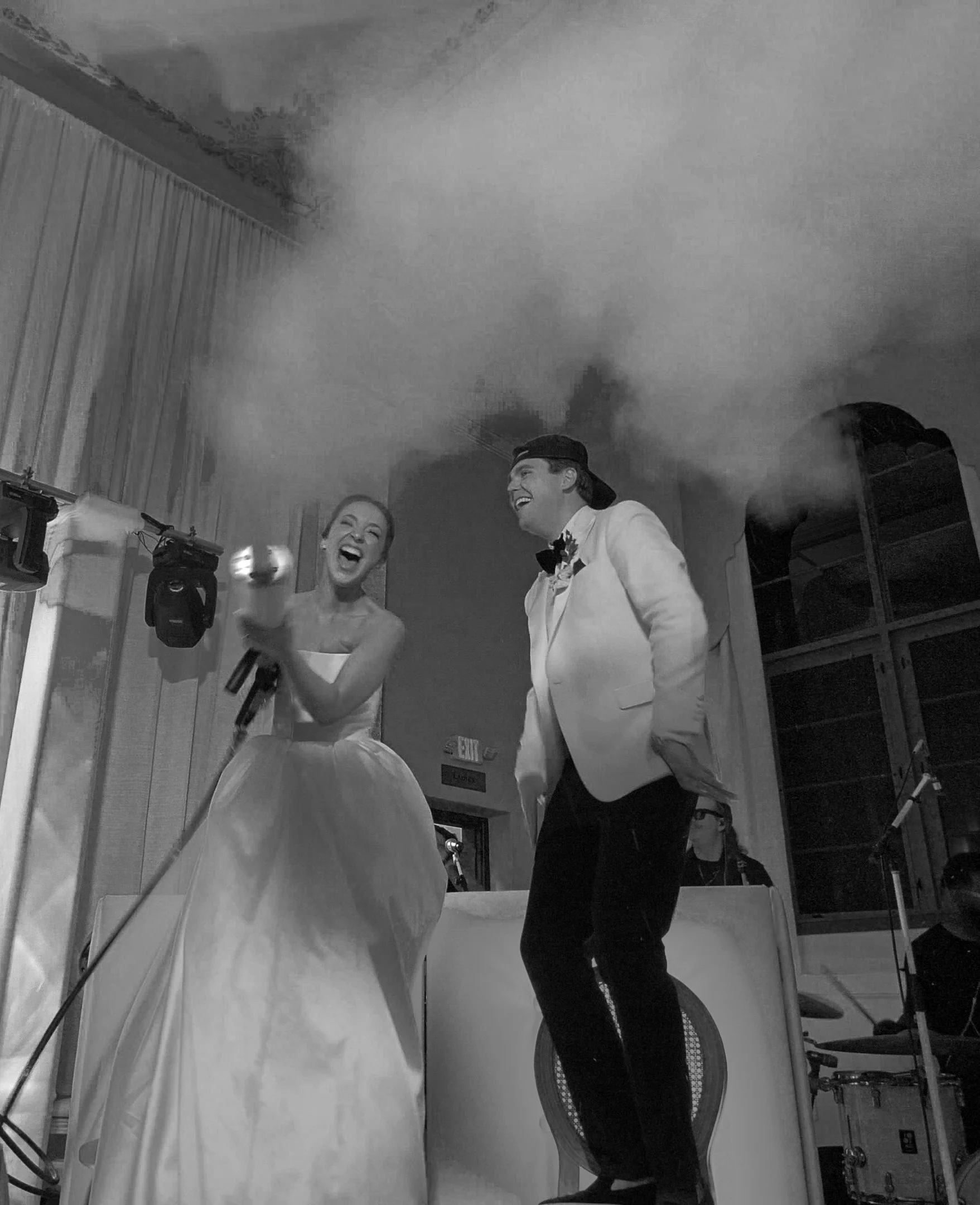 A black and white photo of a happy couple wearing formal attire, standing in front of a wedding cake, with a smoke or fog-like effect in the background.