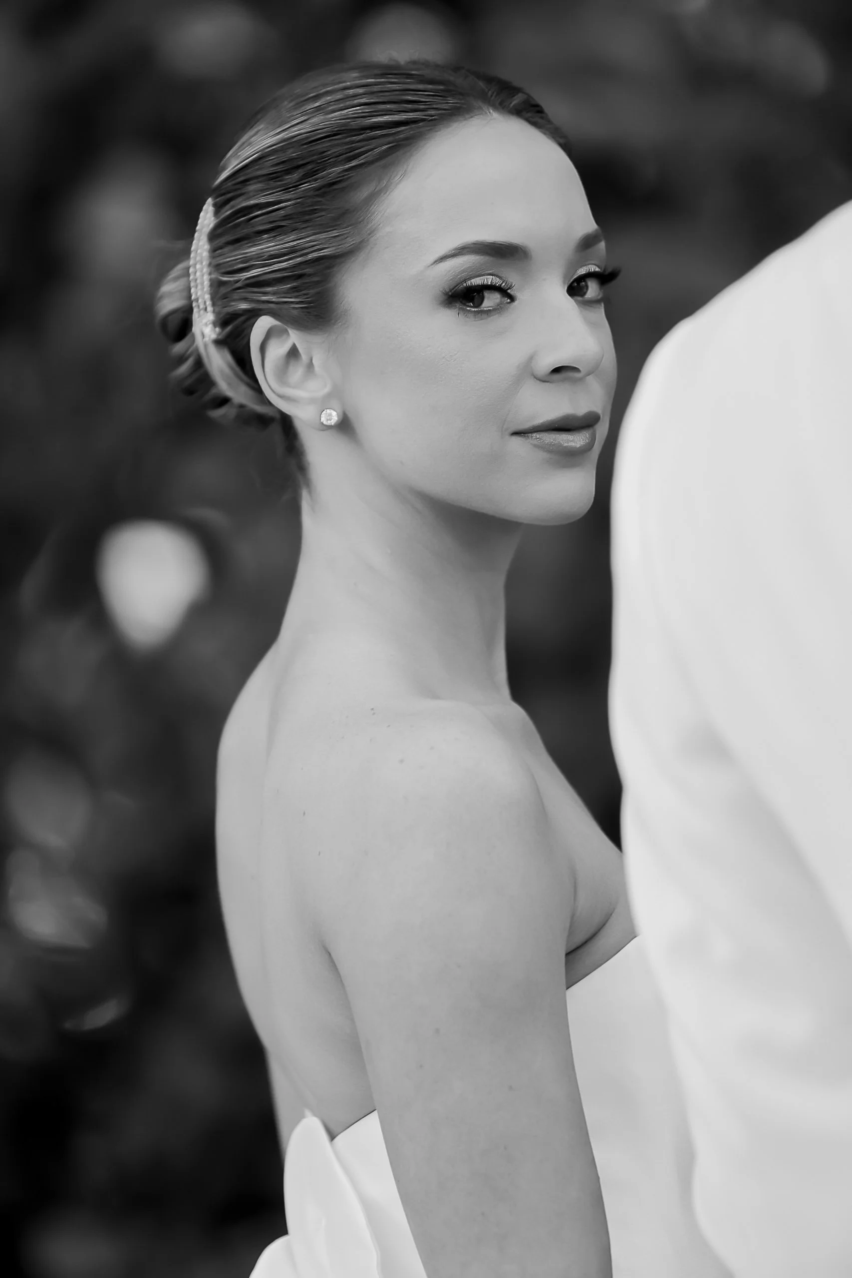 A woman with tied-back hair, wearing earrings and makeup, looking over her shoulder at the camera in a formal setting.
