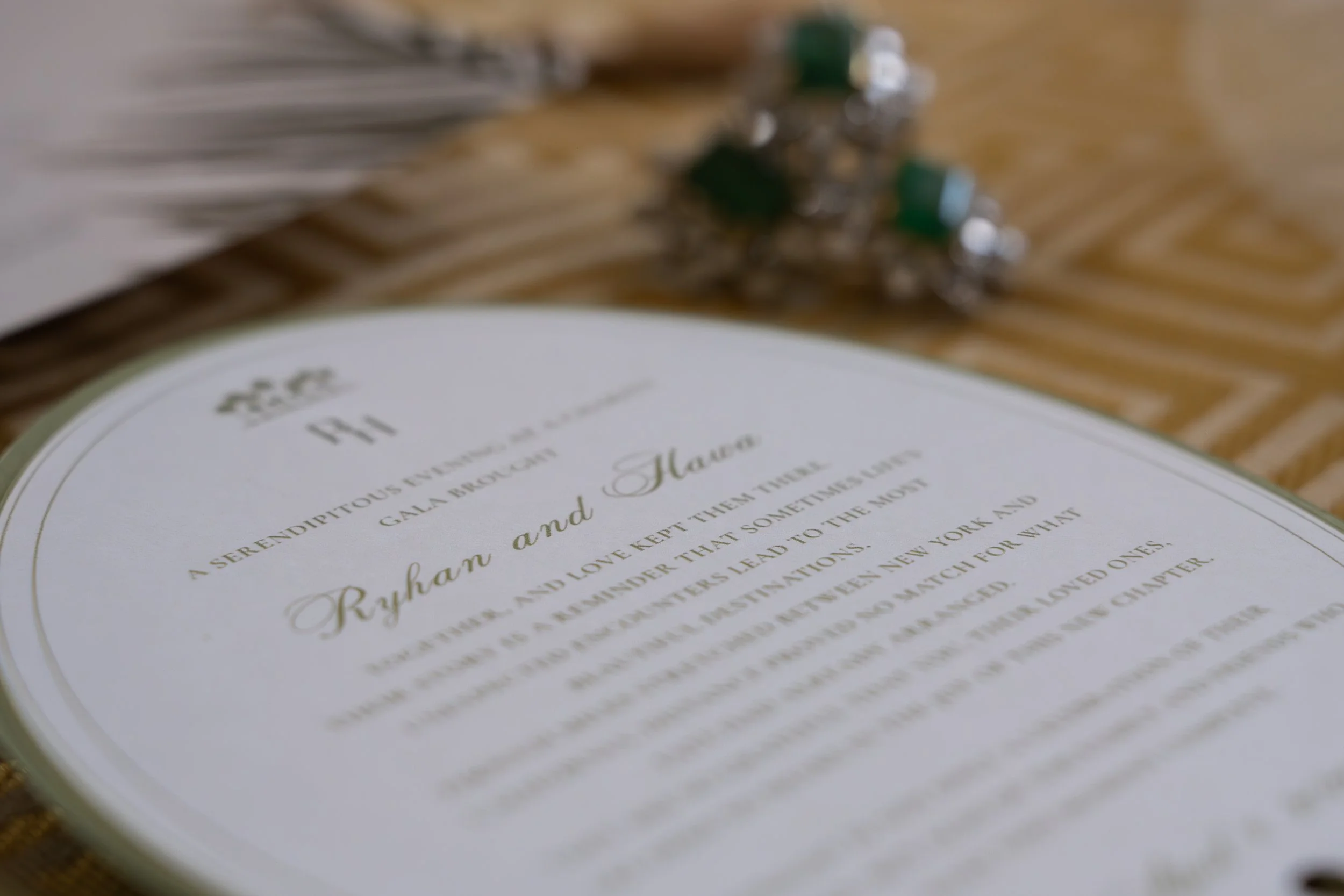 Close-up of a wedding invitation with elegant cursive and serif fonts, placed on a wooden surface with a blurred small bouquet of flowers in the background.