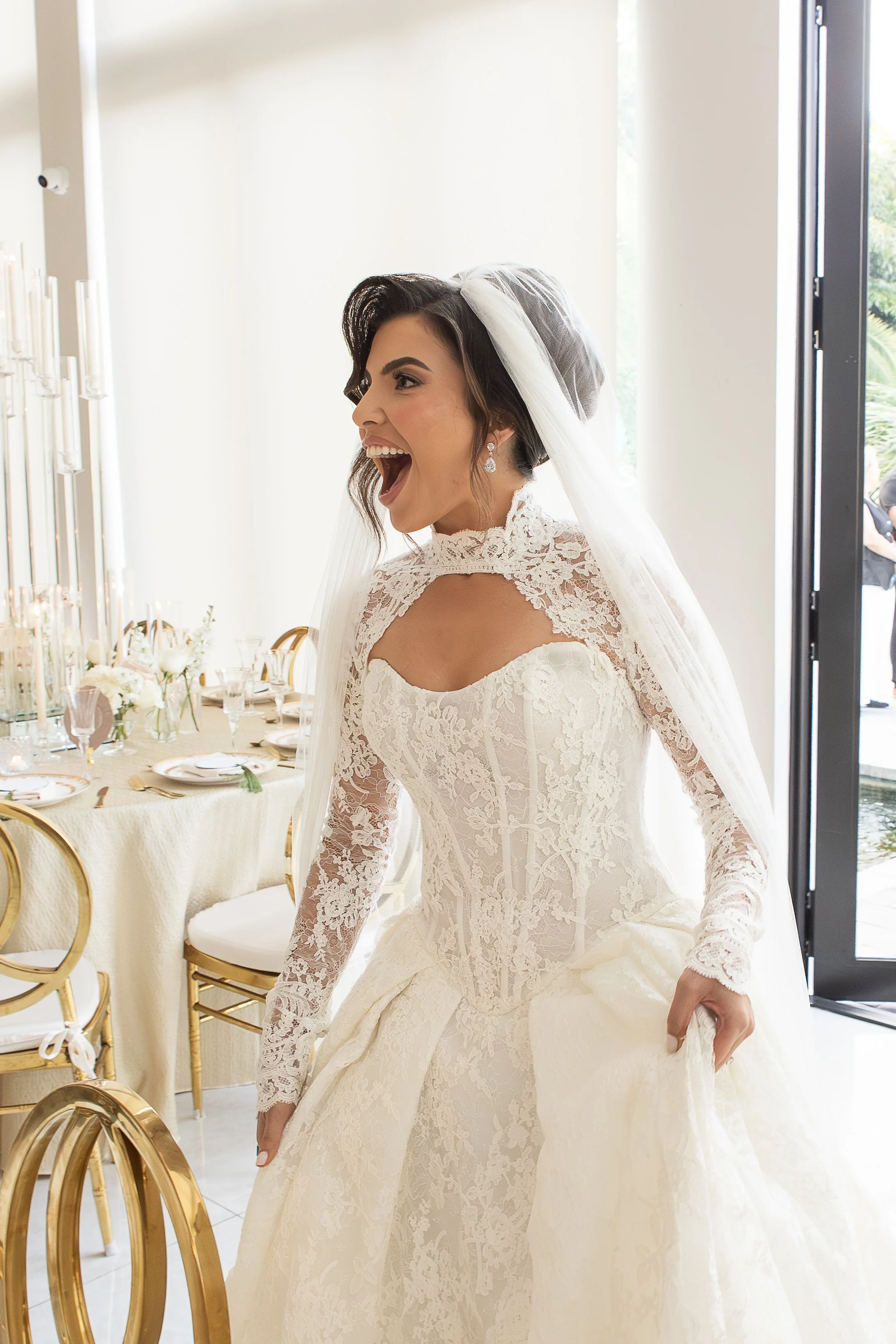 A bride in a lace wedding dress and veil, smiling with her mouth open, inside a bright room with decorated tables and gold chairs.