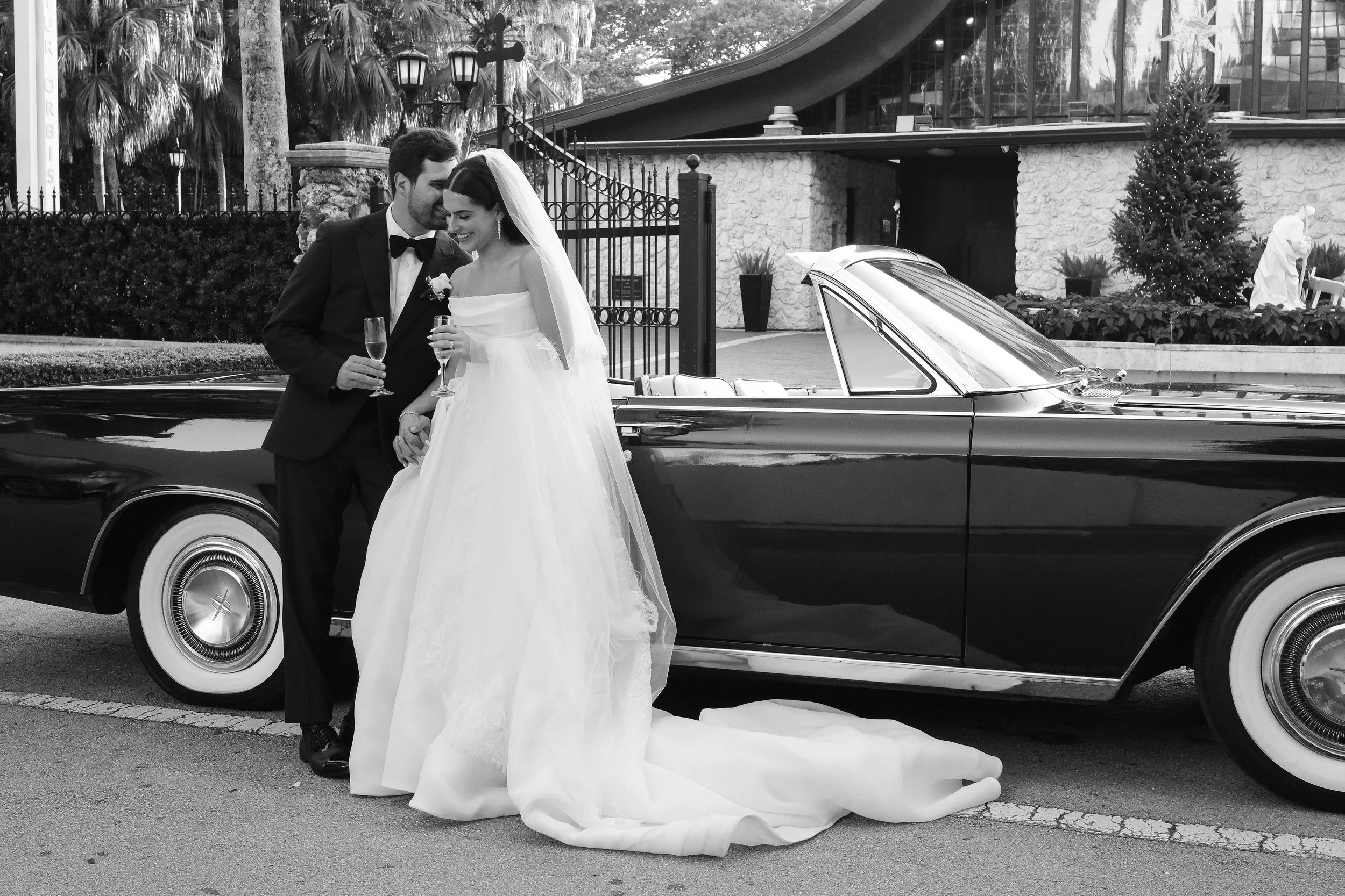 Black and white photo of a newlywed couple standing next to a classic car, smiling and holding champagne glasses, outside in front of a building with trees and a statue in the background.