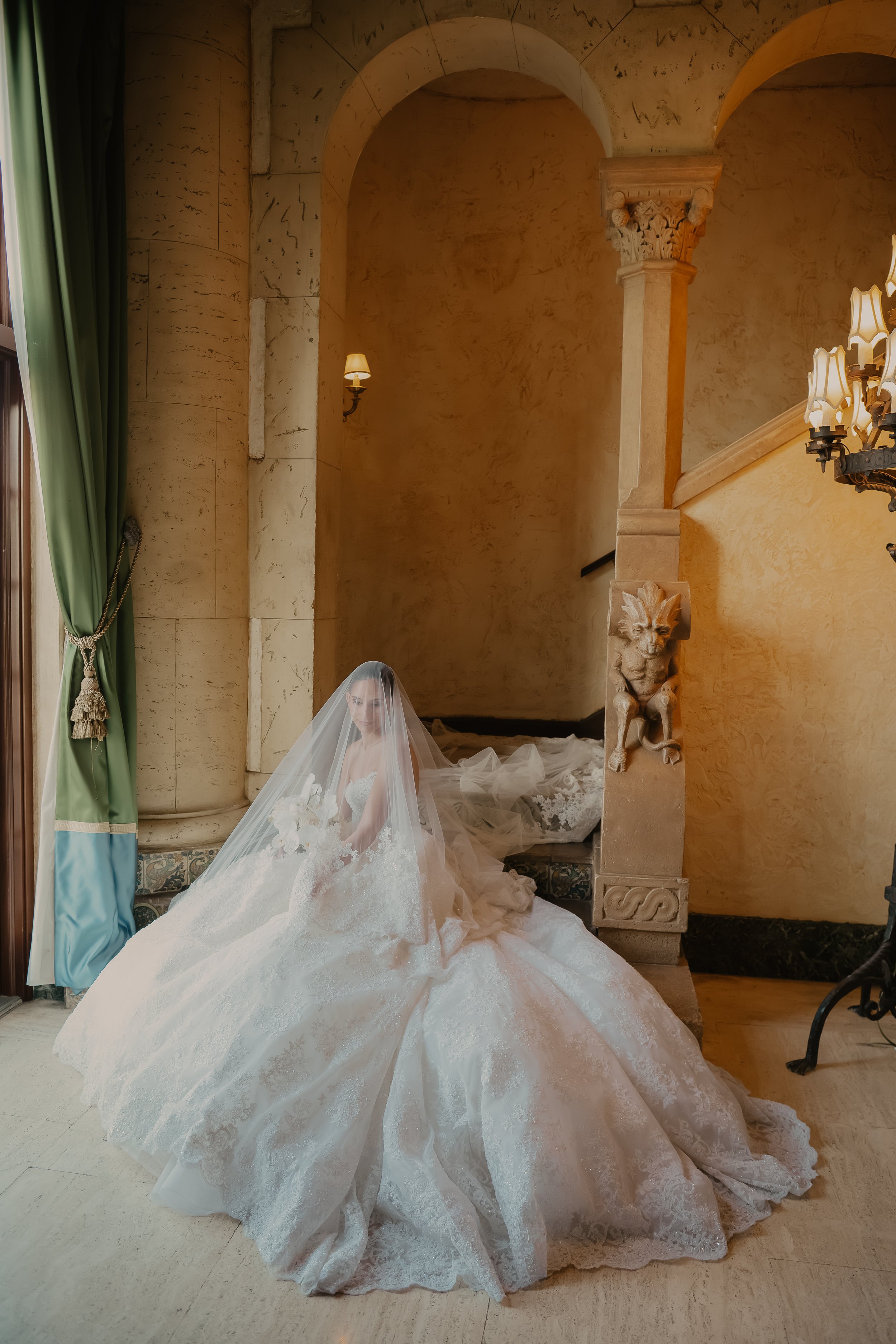 A bride in a wedding dress with a long train and veil sitting on the floor in an elegant, warmly lit room with classical decor, including a lion sculpture on a pillar and a chandelier.
