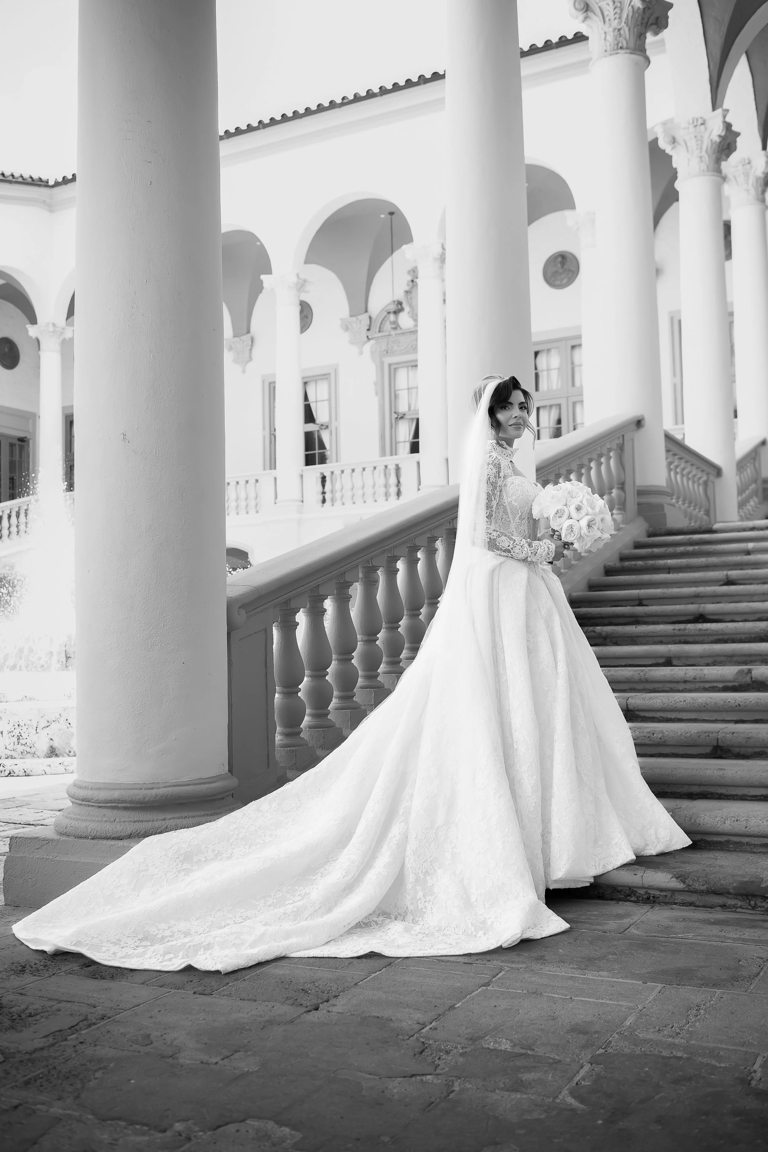 A bride in a white wedding dress holding a bouquet of roses, standing on outdoor stairs in front of a classical building with large columns and arches.