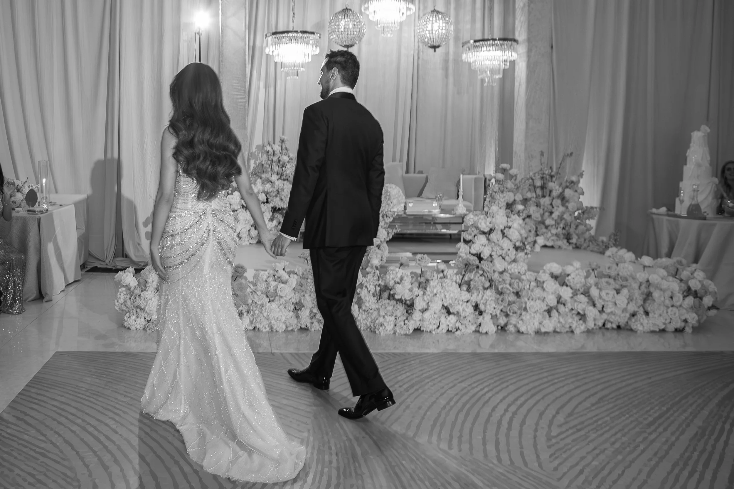 Black-and-white photo of a bride and groom holding hands and dancing at their wedding reception. The bride wears a long, elegant gown with bead embellishments, and the groom is dressed in a dark suit. The background is decorated with multiple chandel