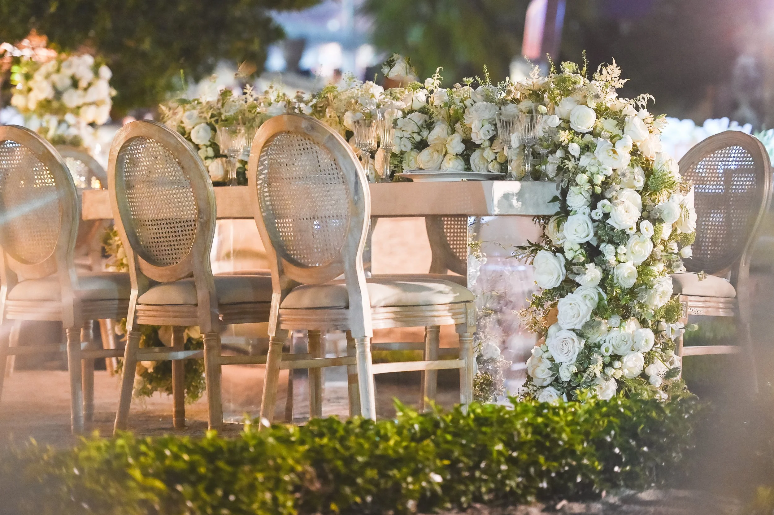 A beautifully decorated outdoor event table adorned with white roses and greenery, with chairs set around it, illuminated with soft lighting in a natural setting.