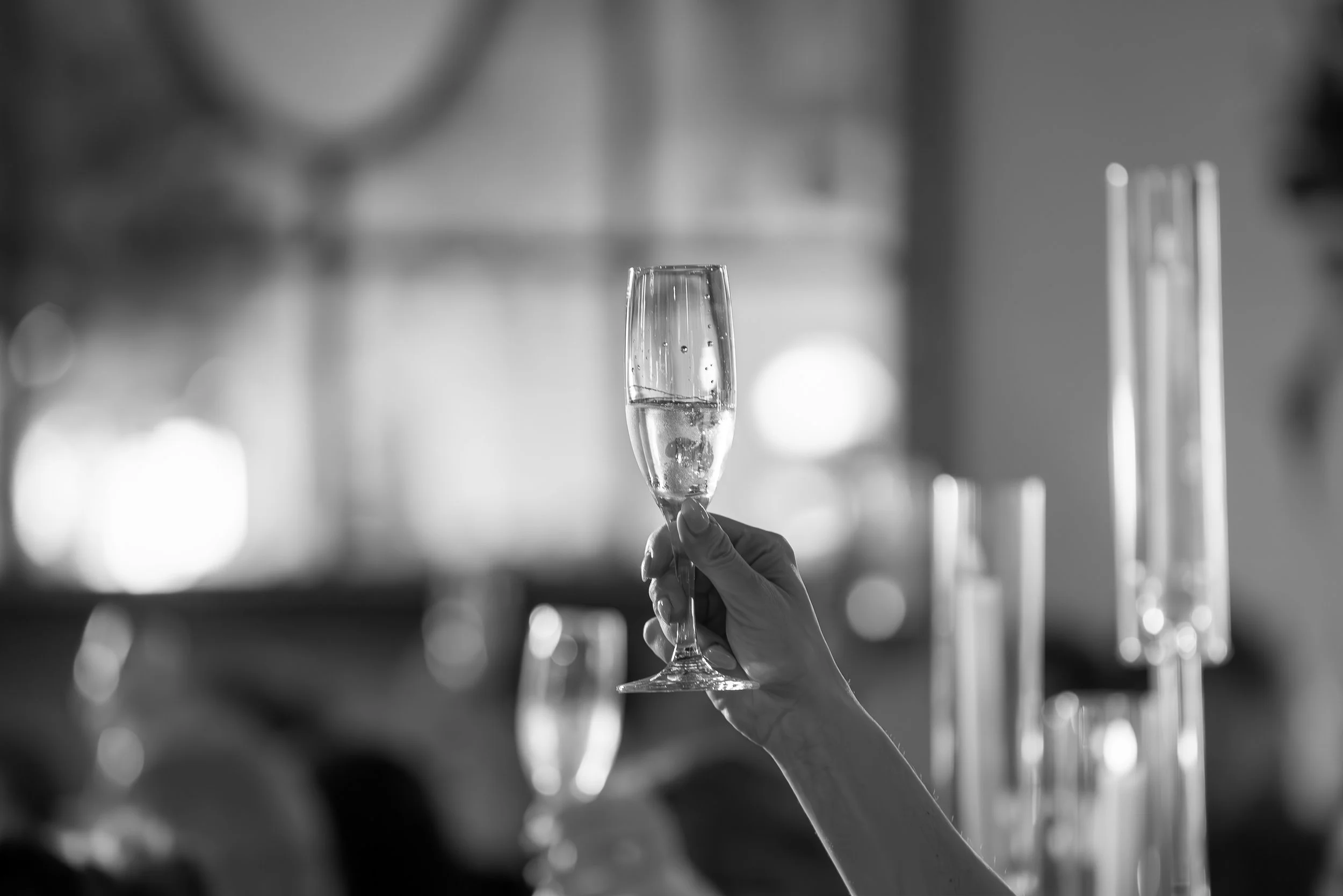 A person holding a champagne flute with a bubbly drink, raised in a toast, in a celebratory setting with blurred lights in the background.