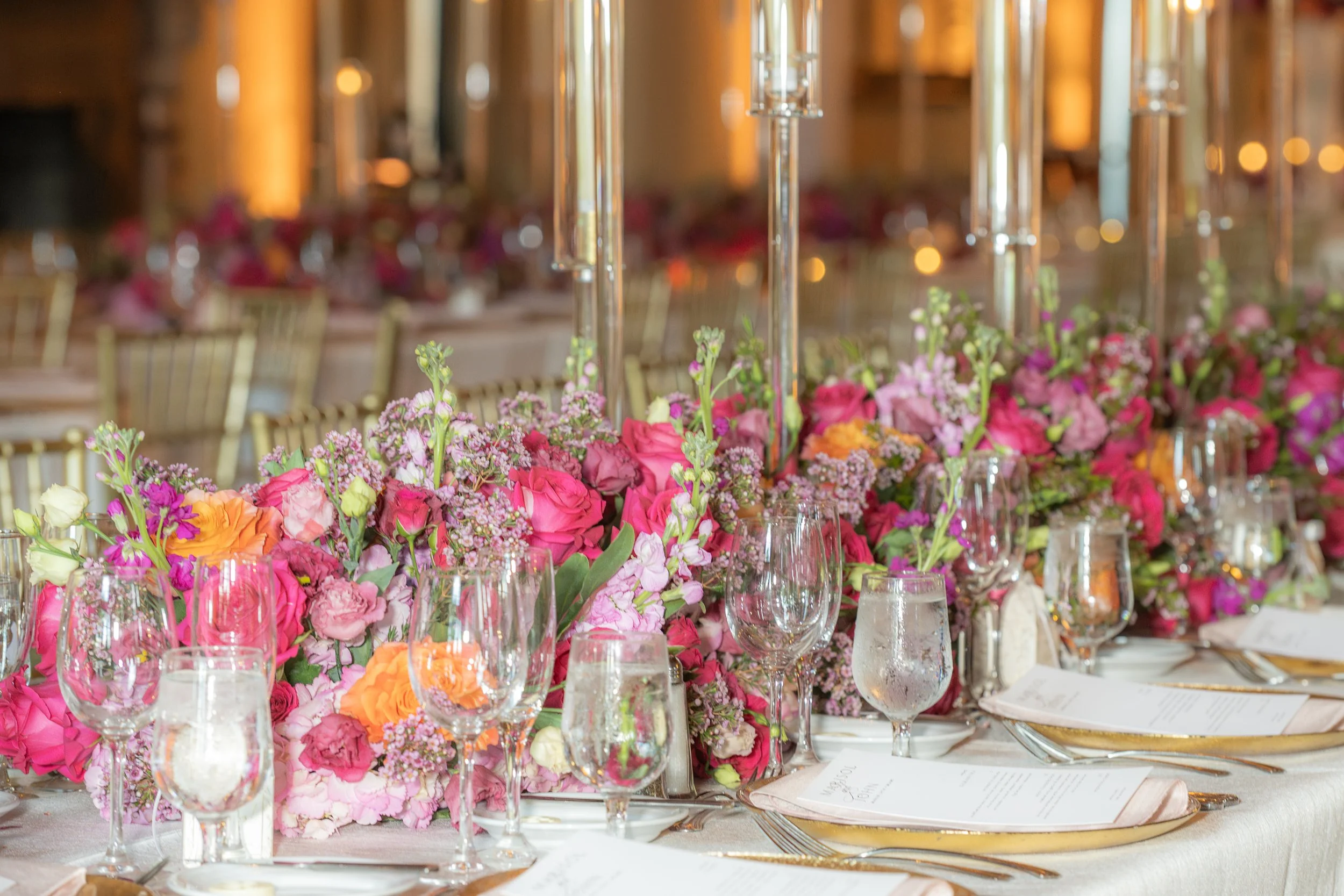 Elegant banquet table with floral centerpieces in pink, purple, and orange, set with wine glasses, water glasses, and gold utensils in a warmly lit room.