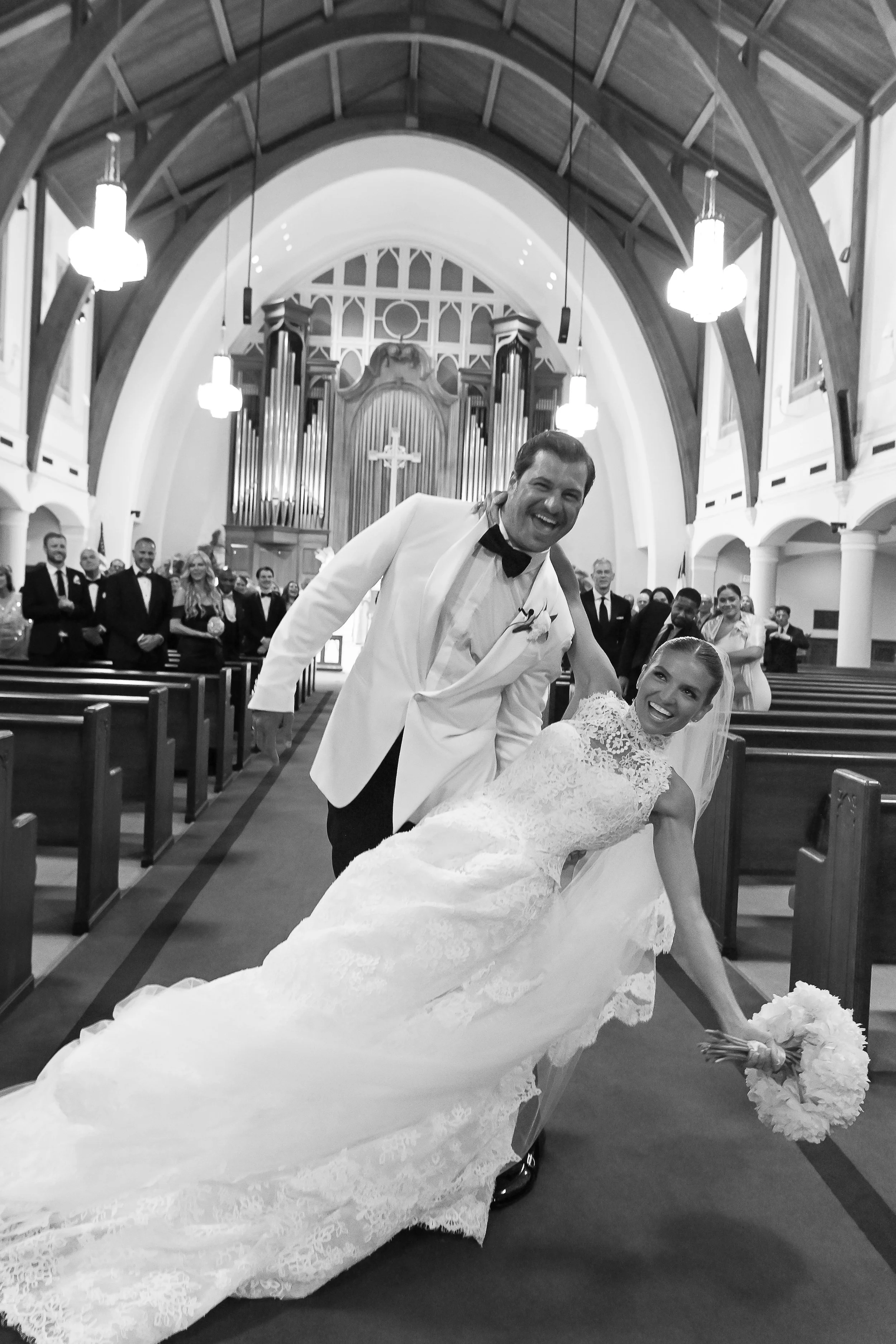 A bride and groom dancing in a church during a wedding ceremony. The bride is leaning back in a dress with a long train, holding a bouquet, and the groom is smiling and leaning over her, wearing a tuxedo. Guests are seated in pews in the background.