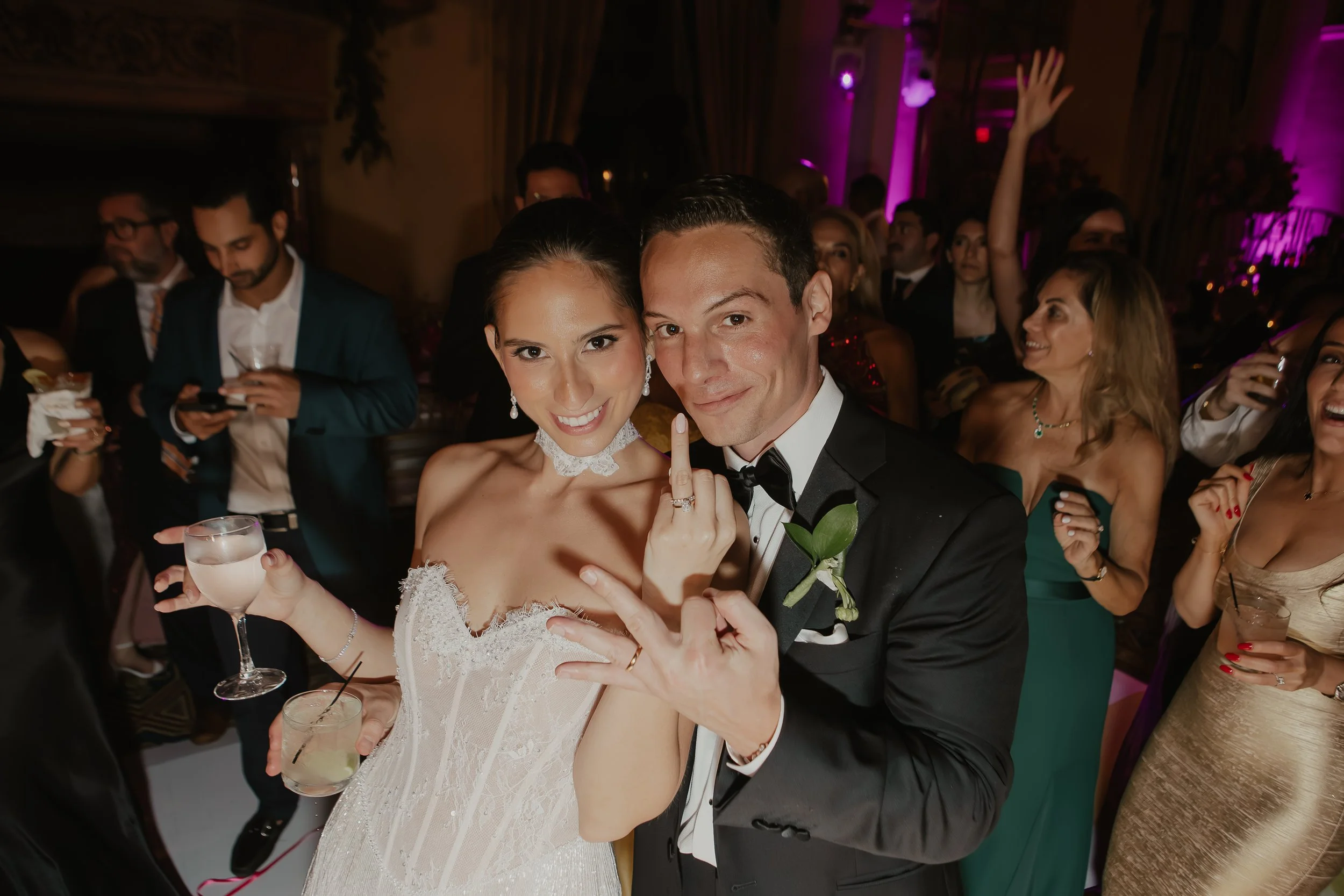 A newlywed couple smiling and celebrating at their wedding reception, surrounded by guests in formal attire, with purple lighting and decorations in the background.
