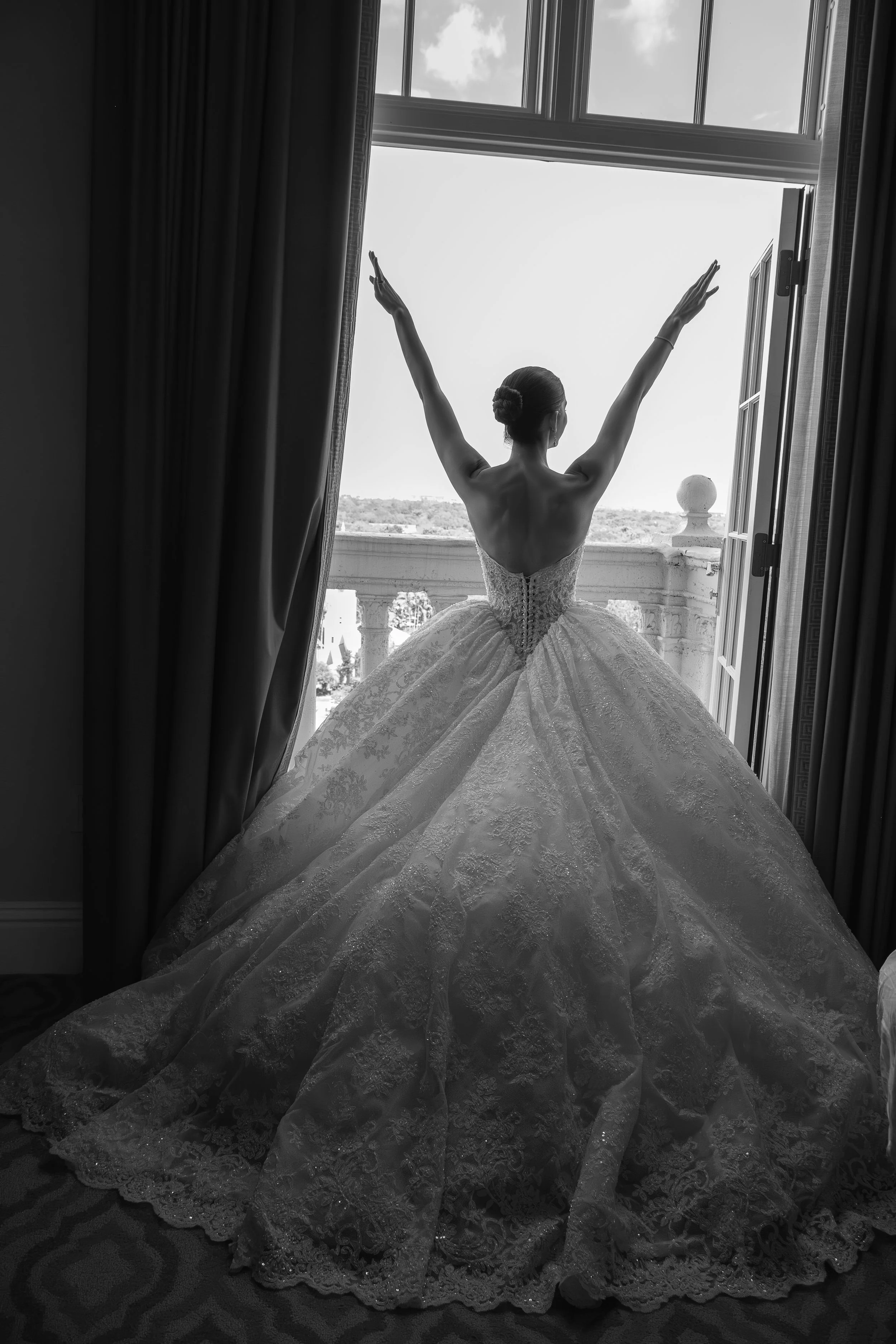 A woman in a wedding dress standing on a balcony, facing away, with arms raised and looking out the open window, overlooking a landscape.