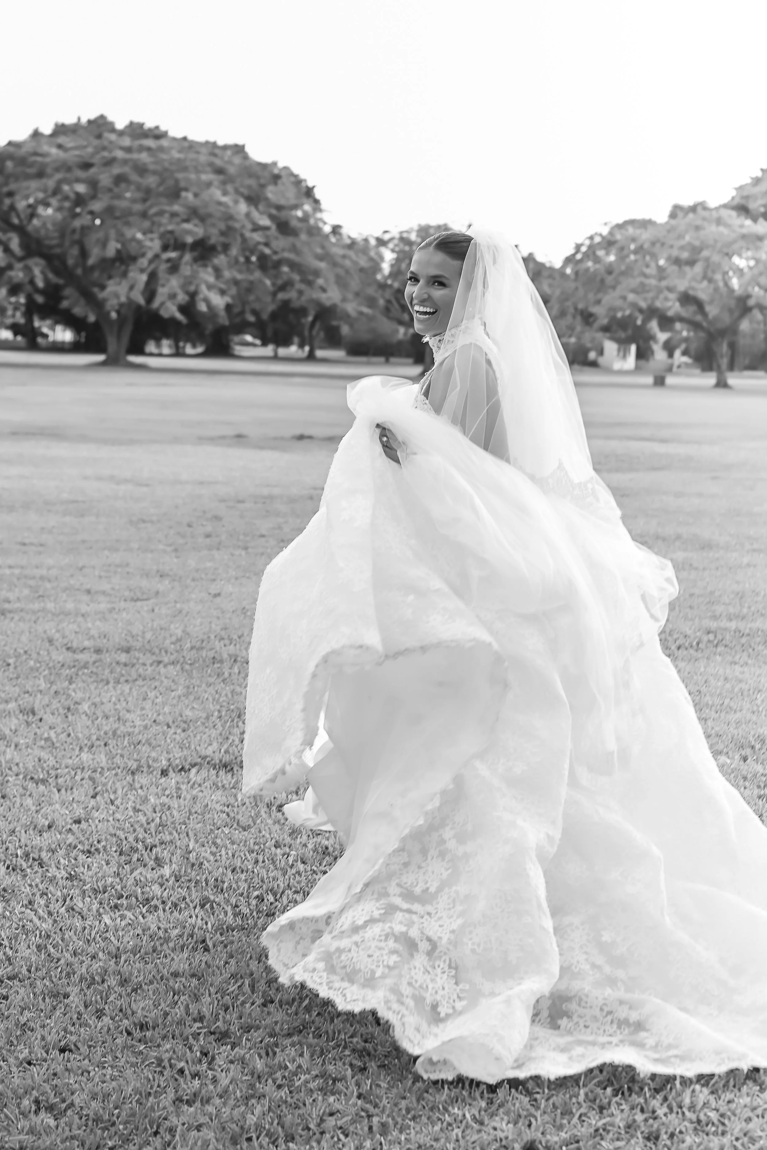 A smiling bride in a wedding dress and veil outdoors on a grassy area with trees in the background.