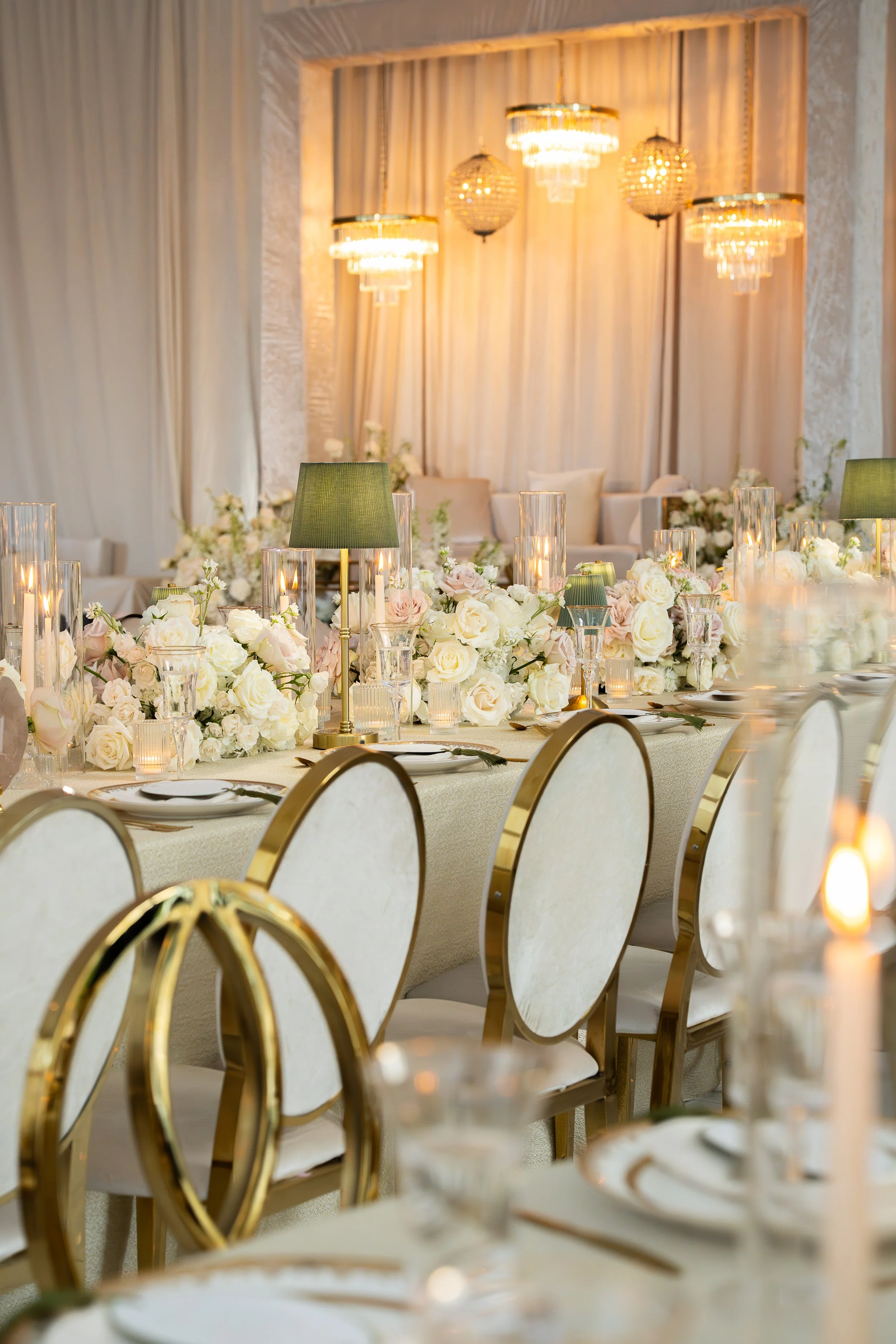 Elegant banquet table decorated with white roses, candles, and green lampshades, set for a formal event in a decorated reception hall with chandeliers and curtains.