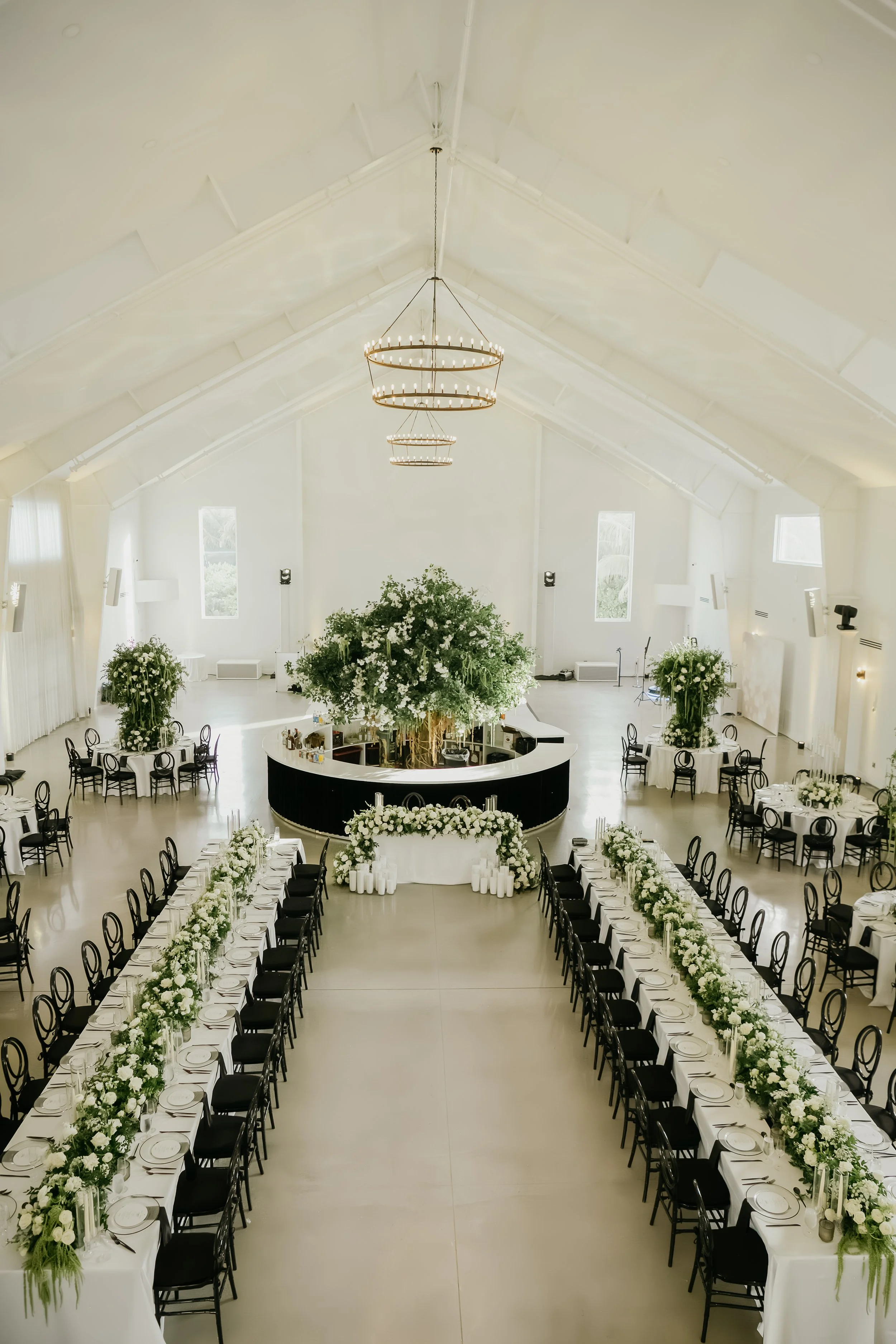 Elegant banquet hall set for a wedding reception with long dining tables decorated with white and green floral arrangements, and a large floral centerpiece at the front stage, with white walls and high ceiling with modern chandelier lighting fixtures