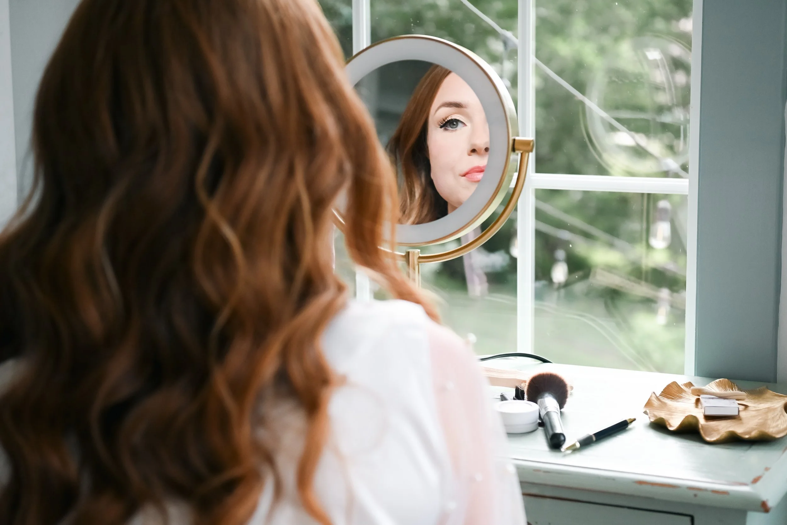 A woman with wavy red hair sitting at a vanity table looking in a round mirror, with makeup tools and brushes on the table, outside greenery visible through the window.