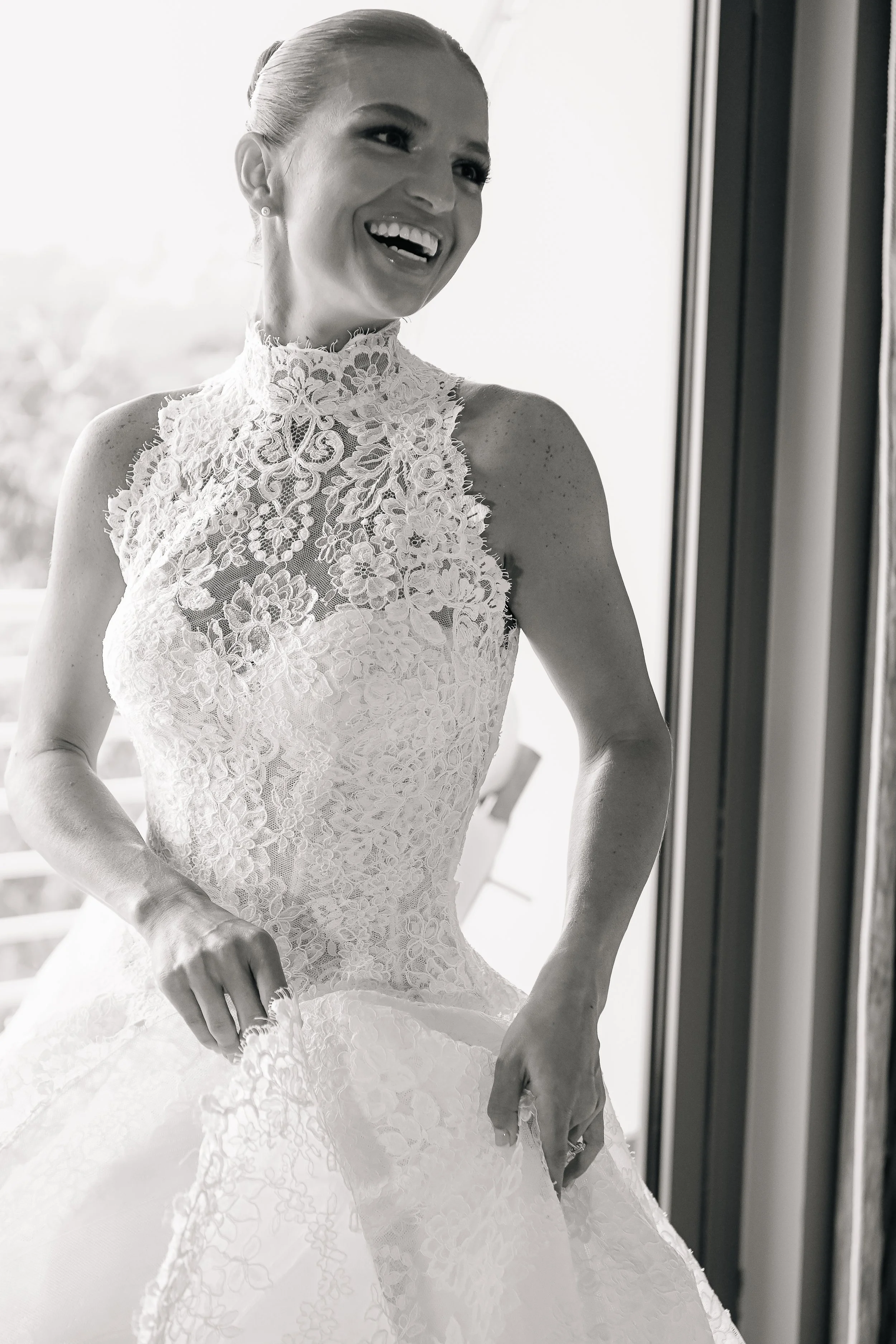 A woman wearing a lace wedding dress standing near a window, smiling and looking to the side.