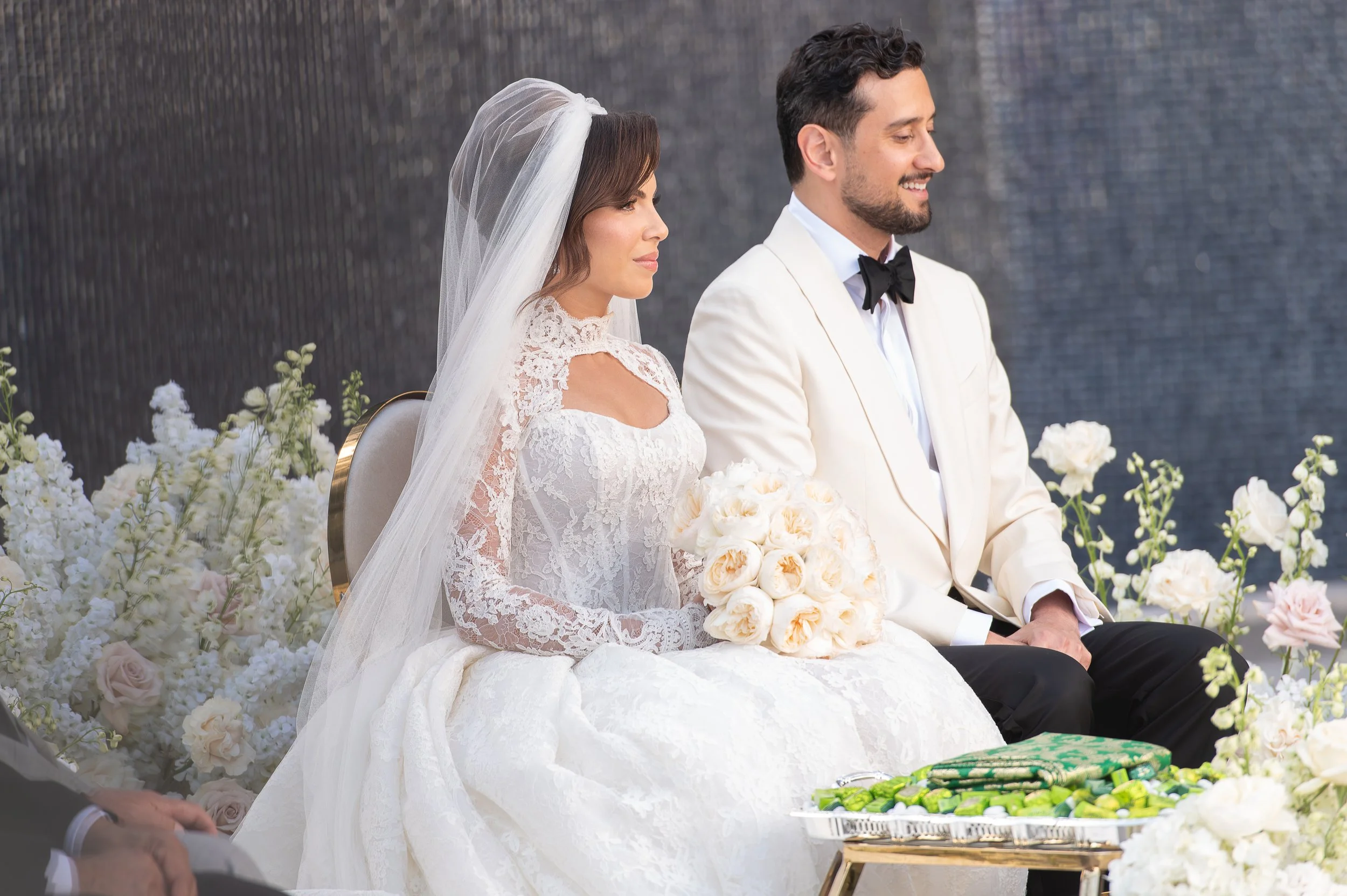 Bride and groom sitting side by side during their wedding ceremony, surrounded by white and pale pink flowers, with the bride wearing a lace wedding dress and veil, and holding a bouquet, while the groom is dressed in a white tuxedo jacket with a bla