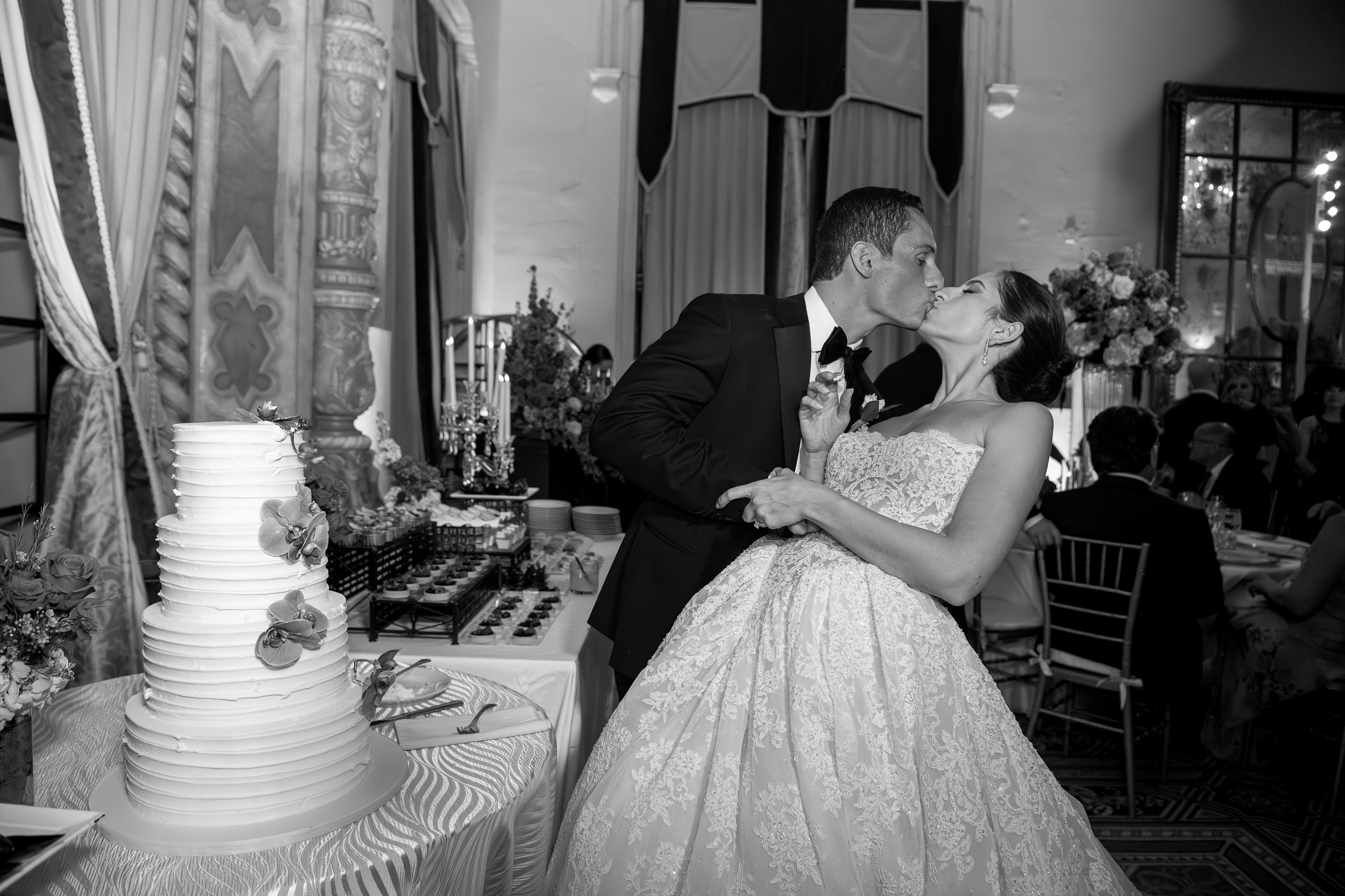 A black and white photo of a bride and groom sharing a kiss at their wedding reception in an ornate decorated ballroom, with a wedding cake and dessert table nearby.