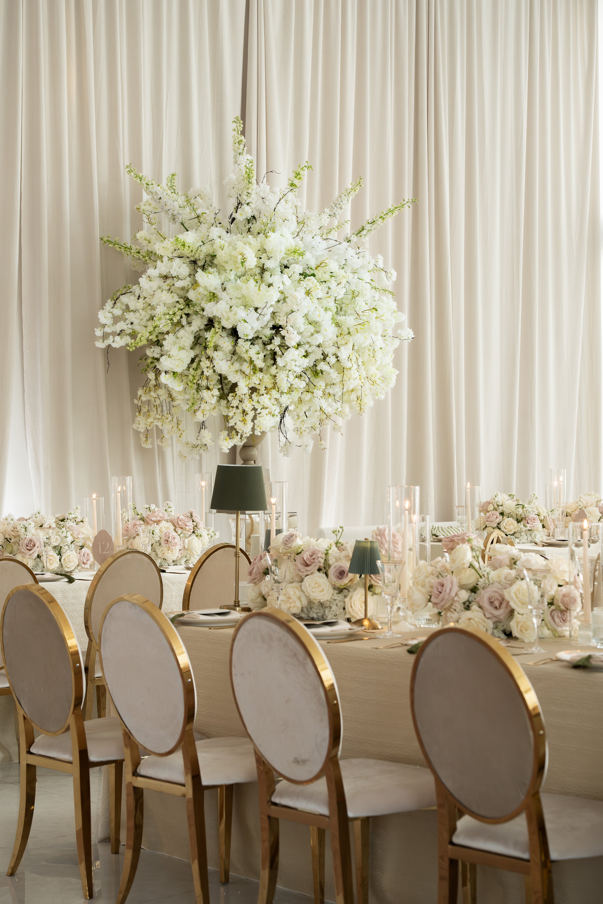 Elegant banquet table decorated with white and blush flowers, tall floral centerpiece with white blossoms, surrounded by gold-rimmed chairs, set for a wedding or special event, with cream-colored curtains in the background.