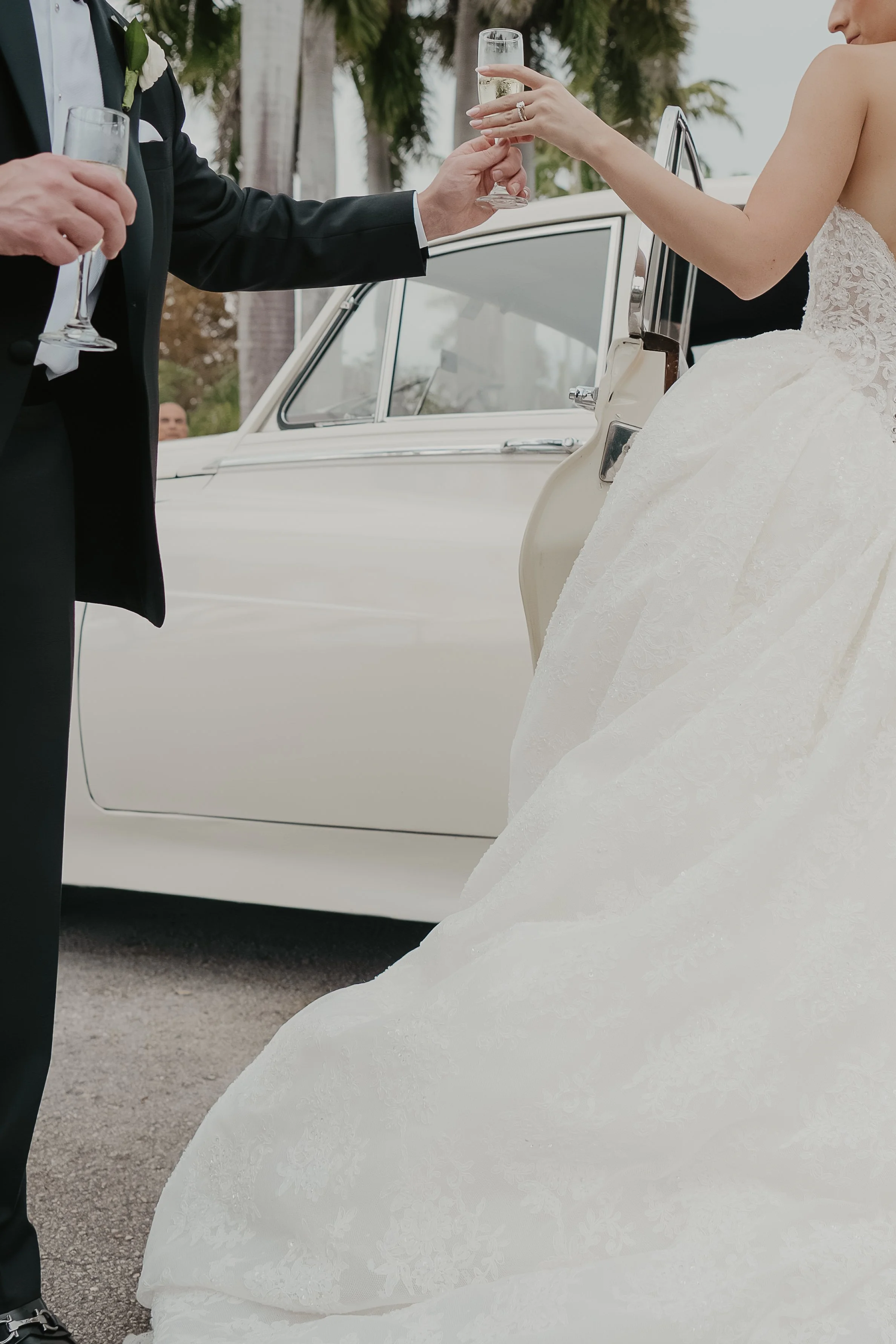 A bride and groom exchanging champagne glasses during a wedding celebration, with the bride in a white lace wedding gown and the groom in a black tuxedo, standing outdoors near a vintage white car, with palm trees in the background.