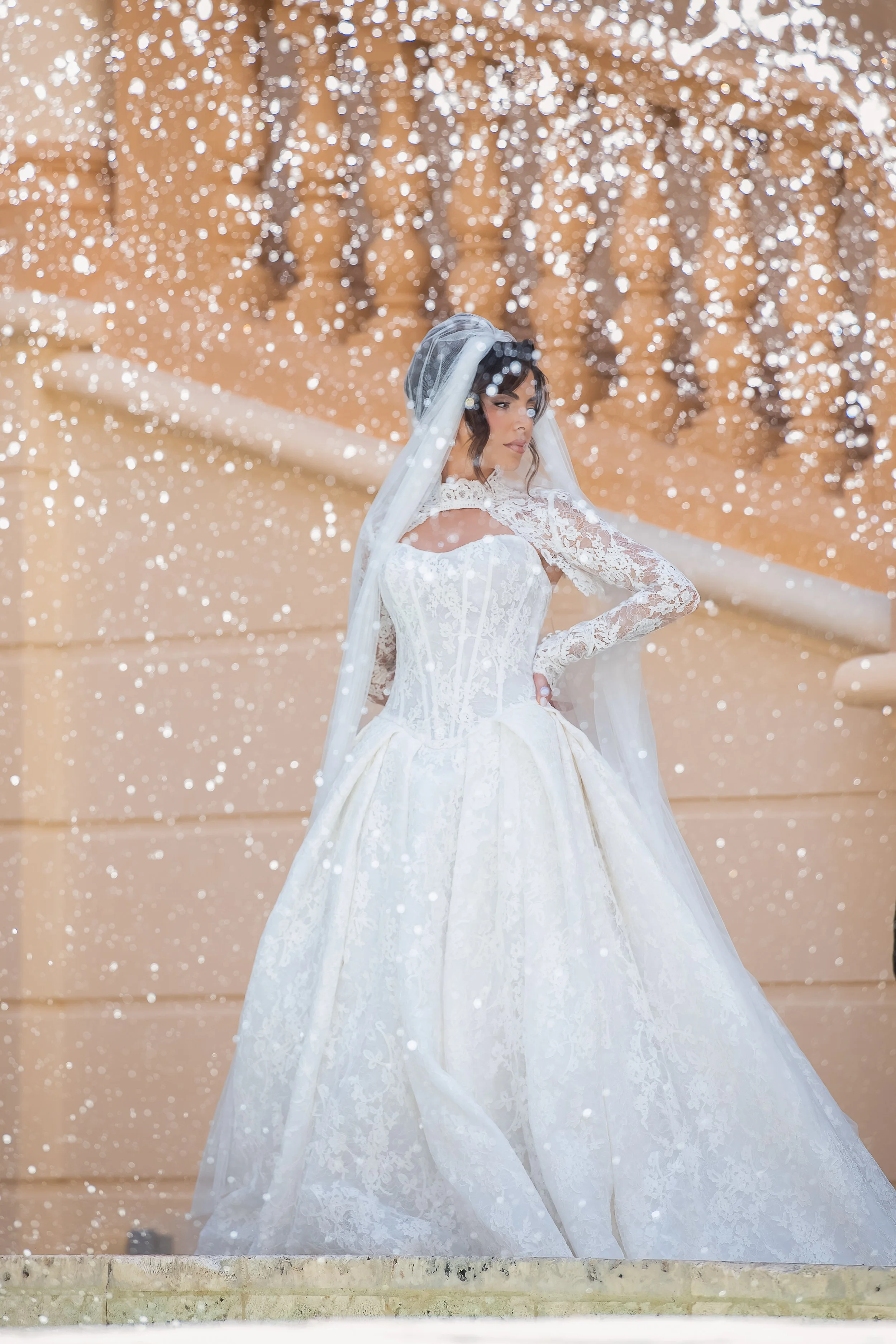 A bride in a white lace wedding gown with long sleeves and a veil stands outside in front of a tan building, surrounded by falling snow.