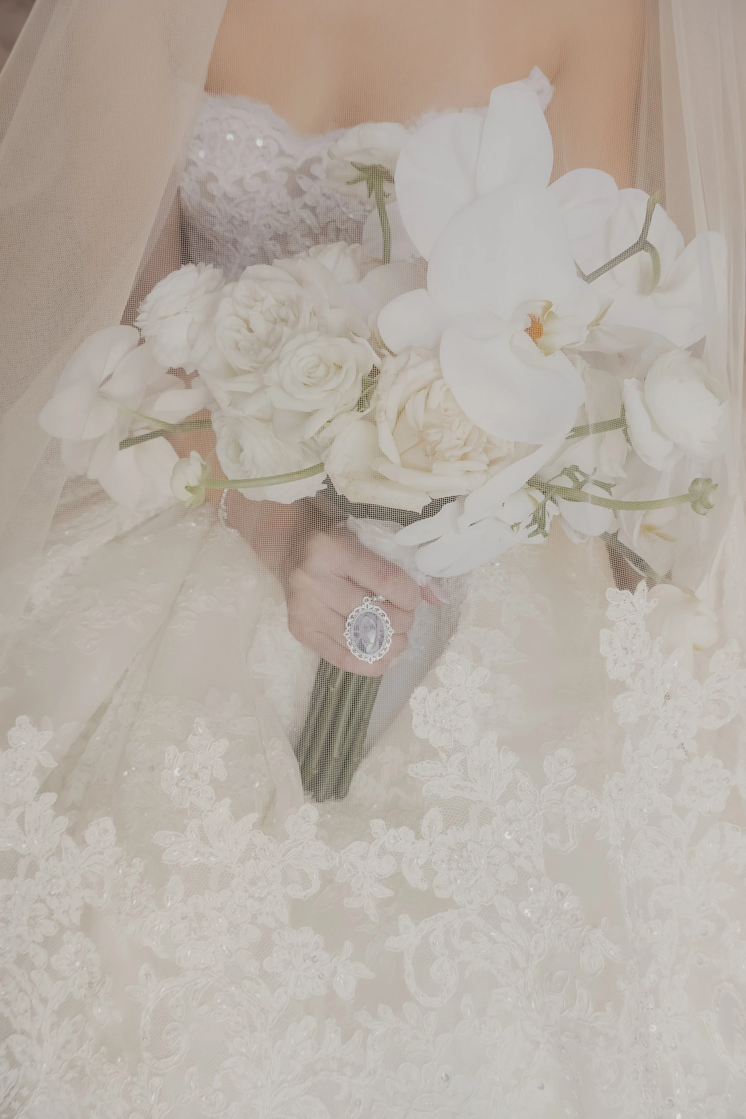 Wedding bouquet with white flowers, including roses and orchids, held behind a wedding veil with lace details.