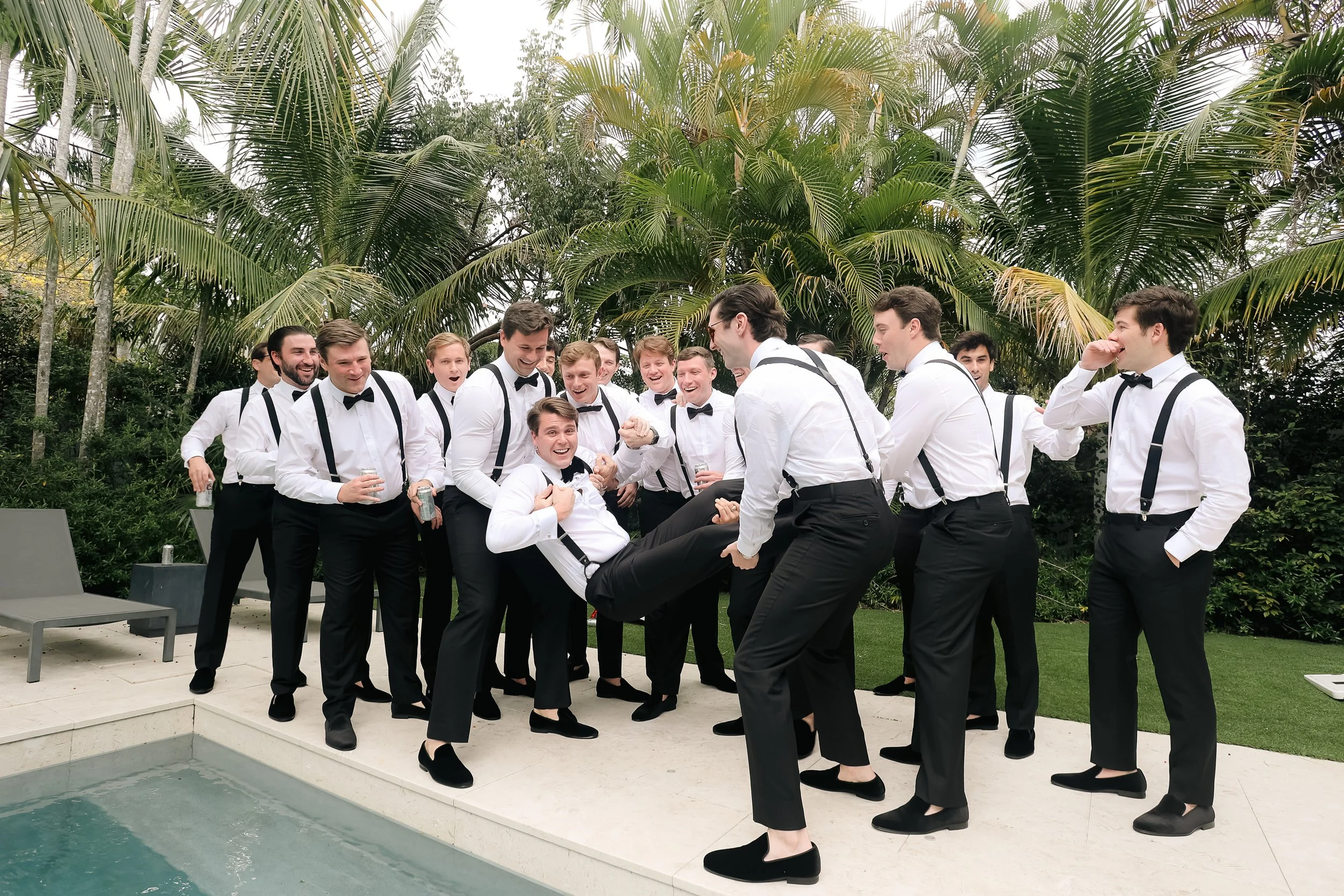 A group of men dressed in white shirts, black pants, suspenders, and bow ties are gathered outdoors near a pool, laughing, smiling, and playfully interacting with each other, with a background of lush green palm trees.