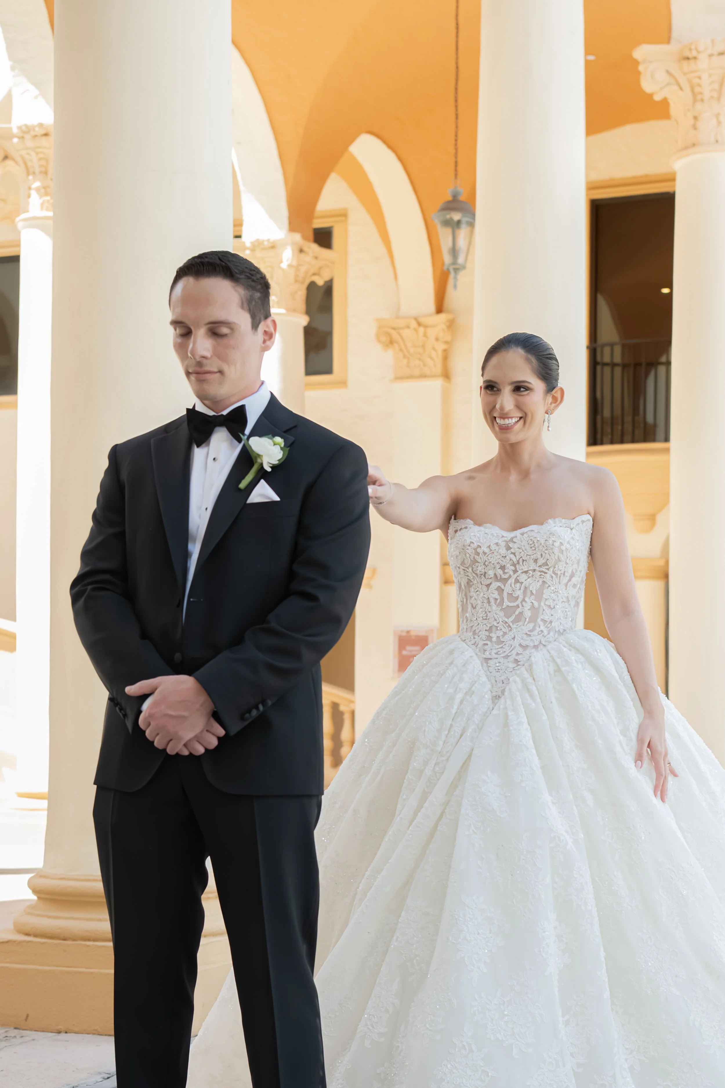 A bride and groom standing inside a building with tall white columns and warm-colored ceilings. The groom is wearing a black tuxedo with a bow tie and a white boutonniere, looking down with his hands clasped. The bride, smiling happily, is wearing a 