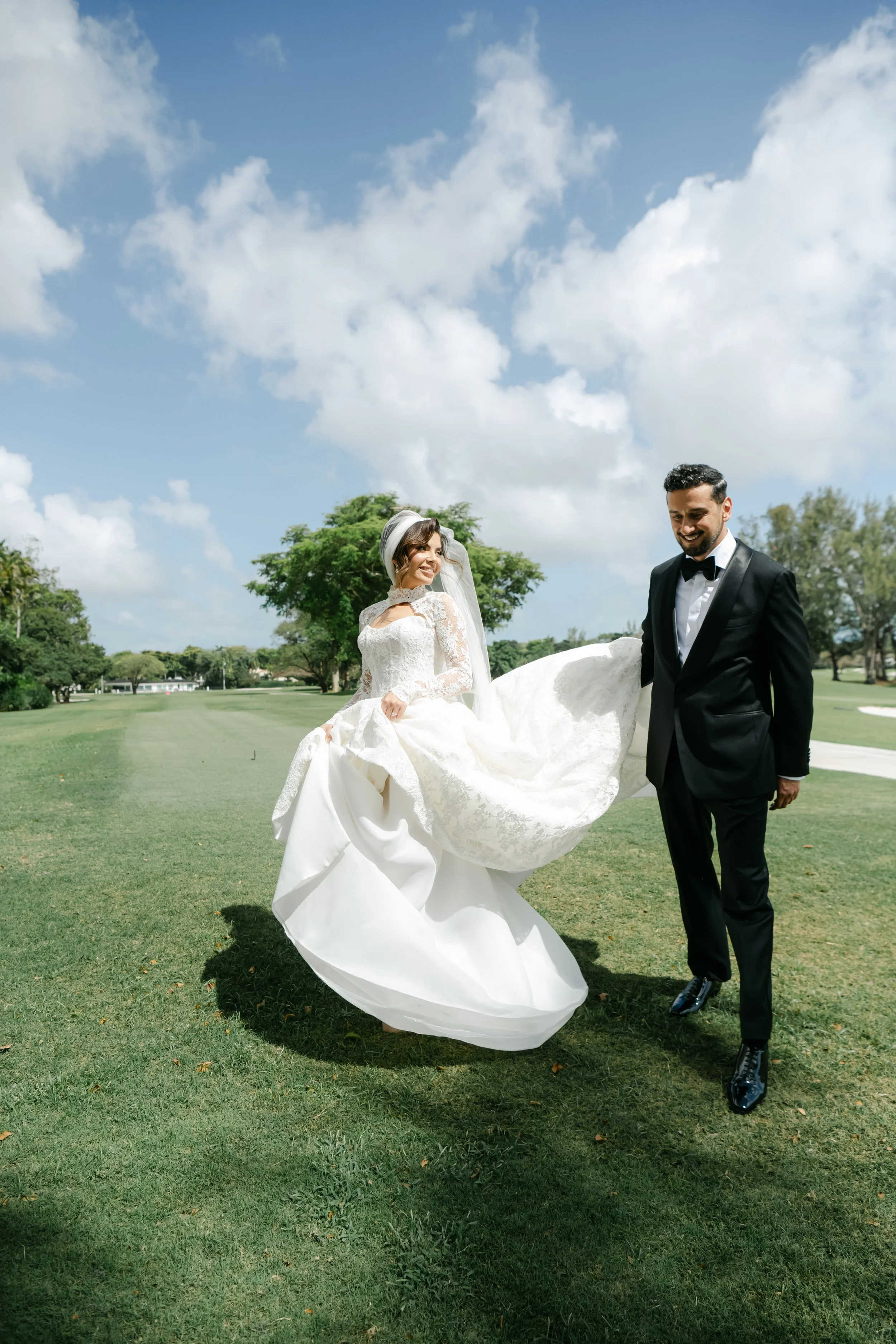 A bride in a white wedding dress with lace details holding her dress up as she walks on grass. A groom in a black tuxedo and bow tie walking beside her, smiling. The background shows green trees and a partly cloudy sky.