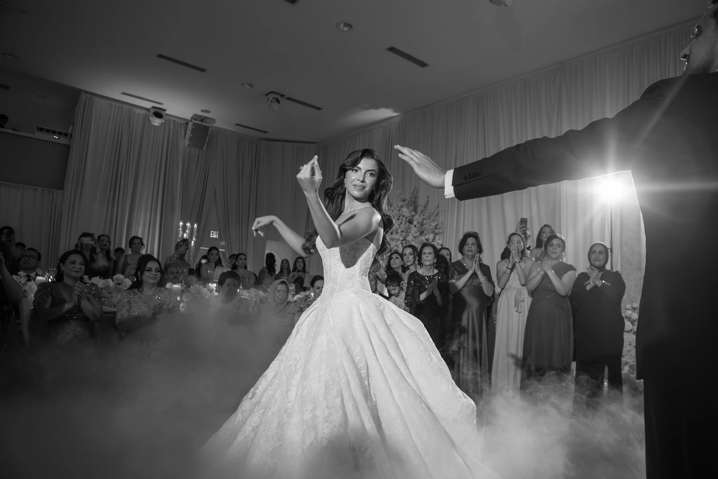 A bride in a wedding gown dancing with a groom during a wedding reception, surrounded by guests clapping and watching inside a decorated hall.