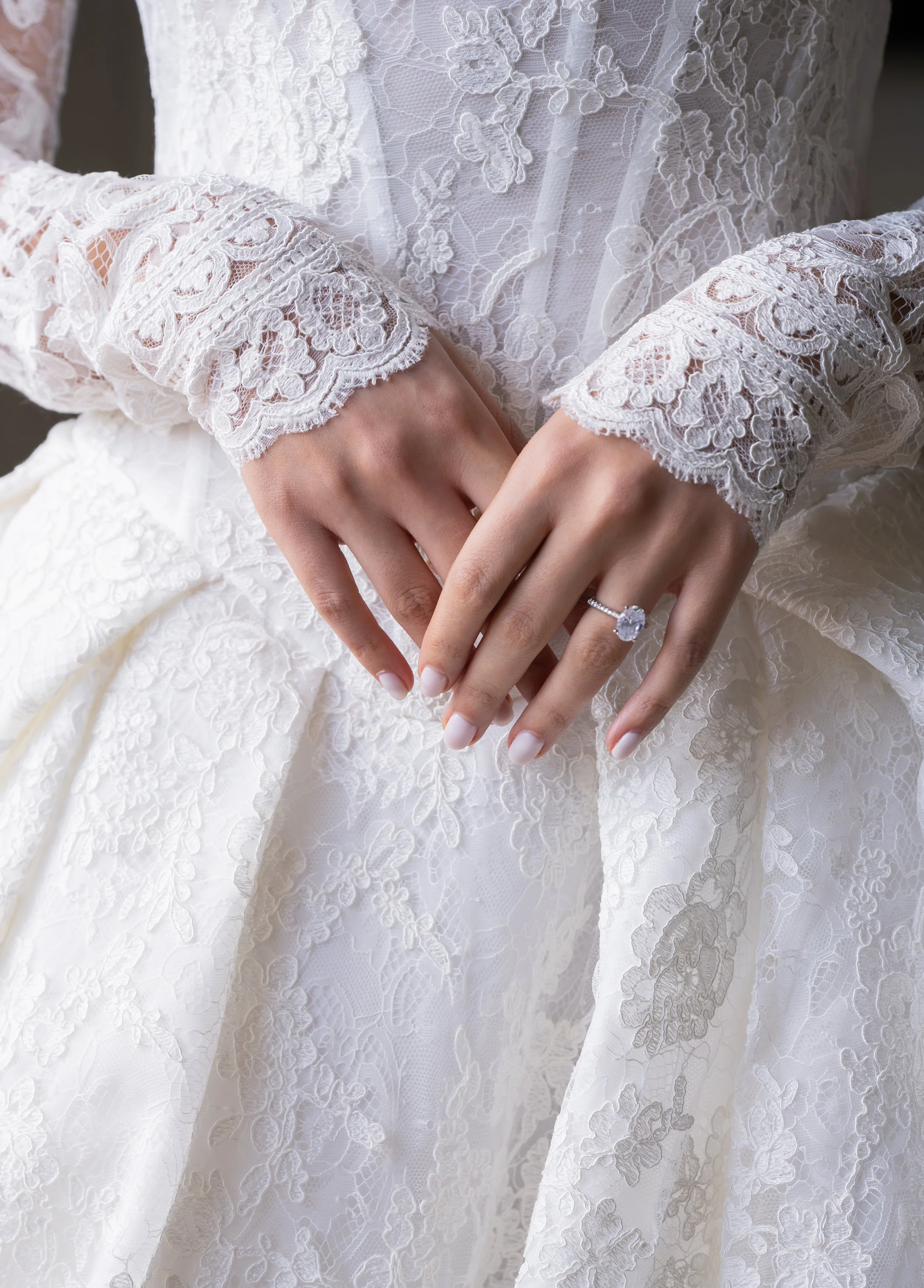 Close-up of a person's hands in a white lace wedding dress, showing a diamond ring on the ring finger of the left hand.