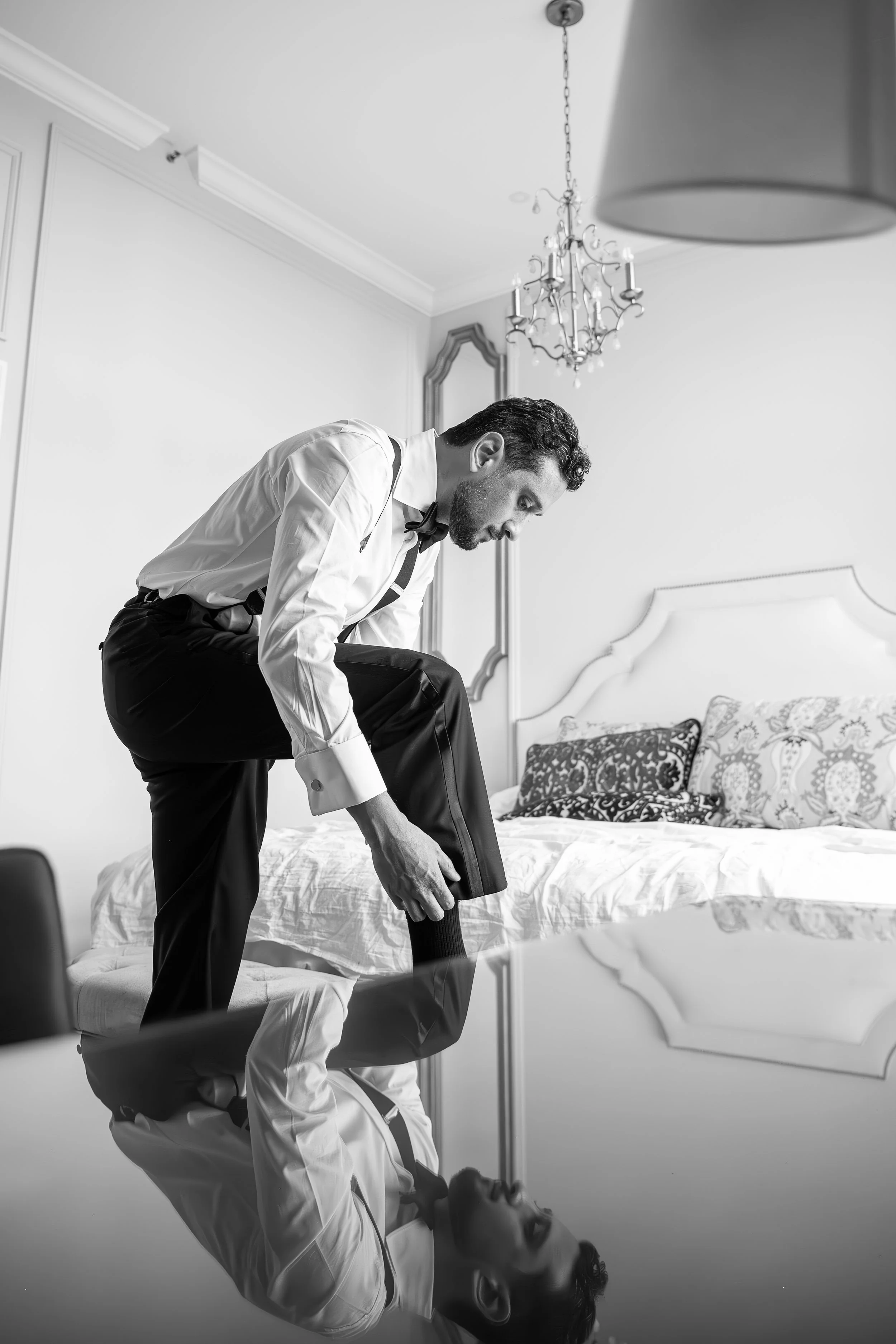 A black and white photo of a groom in tuxedo leaning over a bride lying on a bed, appearing to help her put on her shoes in a bright bedroom with a chandelier and decorative mirror.