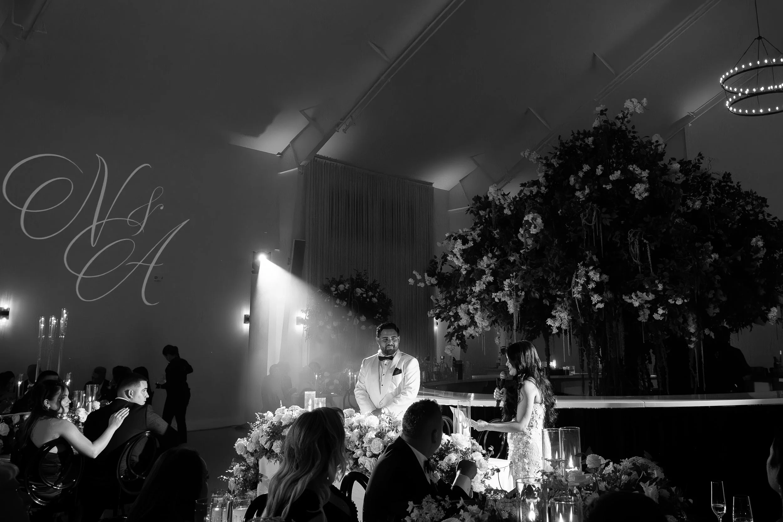 A black-and-white photo of a wedding reception with a man in a tuxedo standing at the head table, surrounded by seated guests, floral arrangements, candles, and a large floral centerpiece. A hanging circular light fixture and a partial view of a chal